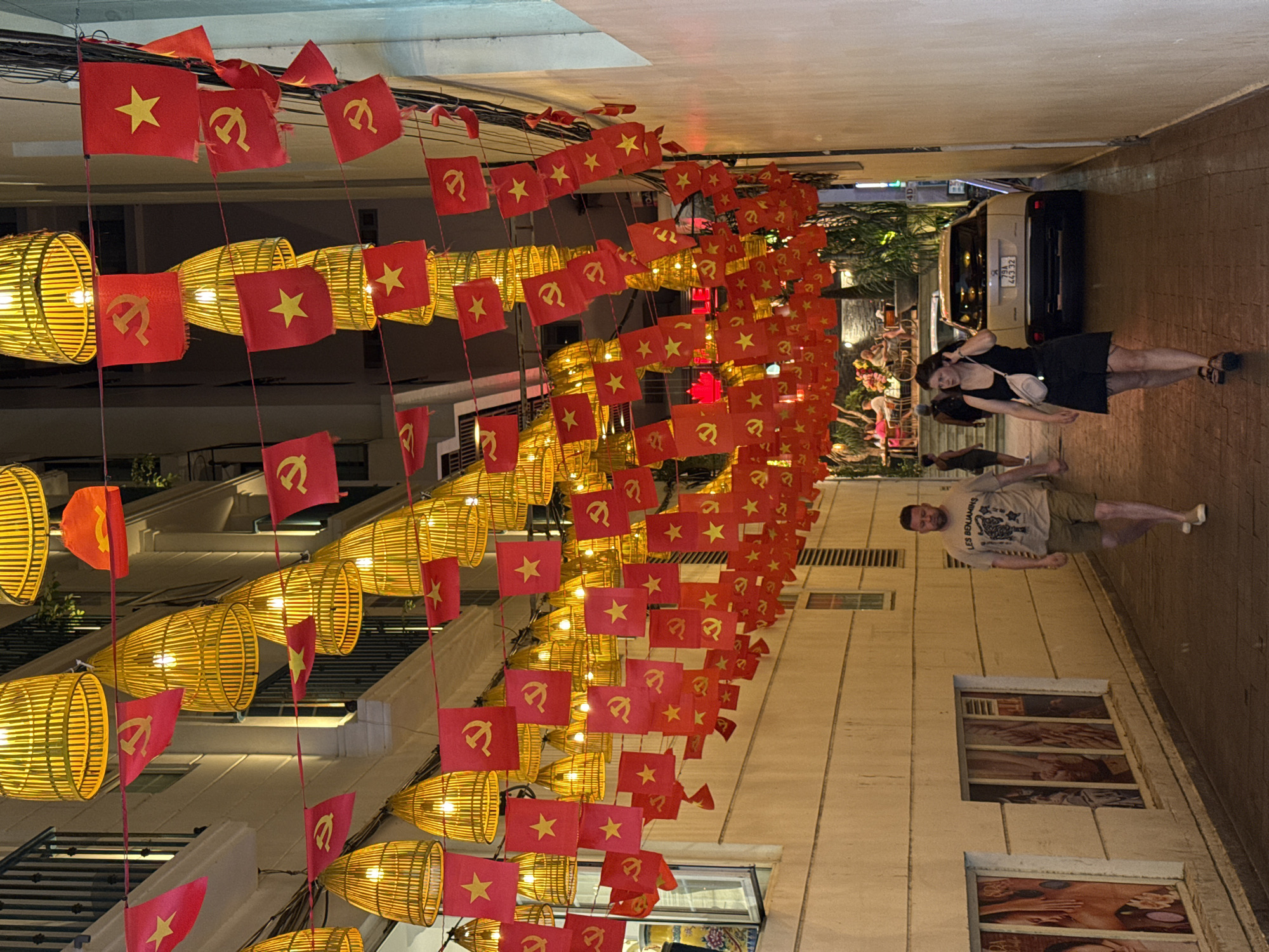 The image depicts a decorated alleyway or narrow street in Nha Trang, Vietnam, captured at what appears to be evening or night. The overhead space is adorned with strings of numerous red flags and yellow, cylindrical bamboo lanterns. The flags alternate between the national flag of Vietnam, featuring a yellow star on a red field, and flags bearing the yellow hammer and sickle symbol on a red field. The lanterns emit a warm, bright light, illuminating the area.
Two people are visible walking on the right side of the street. A woman wearing a dark dress and sandals is walking towards the foreground, looking to her left. She appears to be carrying a small white bag and holding a phone. Further down the street, a man wearing a light-colored t-shirt with "LES BENJAMINS" printed on it and shorts is also walking.
The street is lined with multi-story buildings. On the right, some windows display images, possibly advertisements for a spa or massage parlor, showing hands and faces. In the background, a light-colored car, likely a taxi, is parked; its license plate is visible as "79A 443.32". The scene suggests a lively urban setting, possibly a pedestrian area or a street known for its evening decorations and local businesses.
