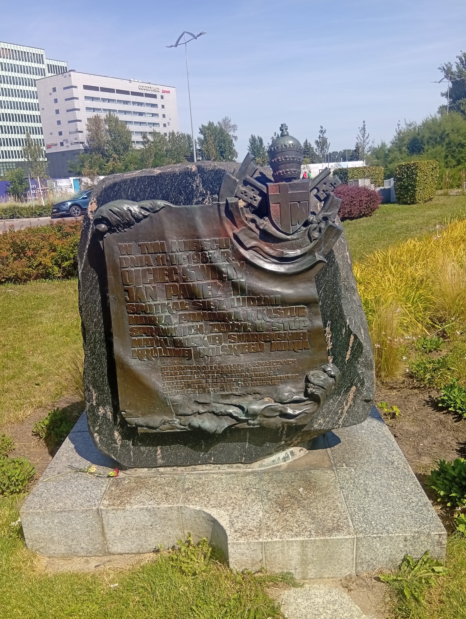 The image depicts a monument located in Gdynia, Poland, specifically at Plac Jana Pawła II. The monument is a large, dark stone boulder with a bronze plaque and a crest. The crest features a crown and crossed keys, symbols associated with religious or papal authority. The bronze plaque contains embossed text in Polish, detailing an event that occurred on June 11, 1987, where Pope John Paul II met with the Kashubian people. The monument is situated in an outdoor setting with a paved base and surrounding greenery, including grass and bushes. In the background, modern buildings, including a Courtyard by Marriott hotel, are visible, suggesting an urban environment. The sky is clear and blue. A single flower lies on the base of the monument. The overall scene conveys a sense of historical commemoration and public space.