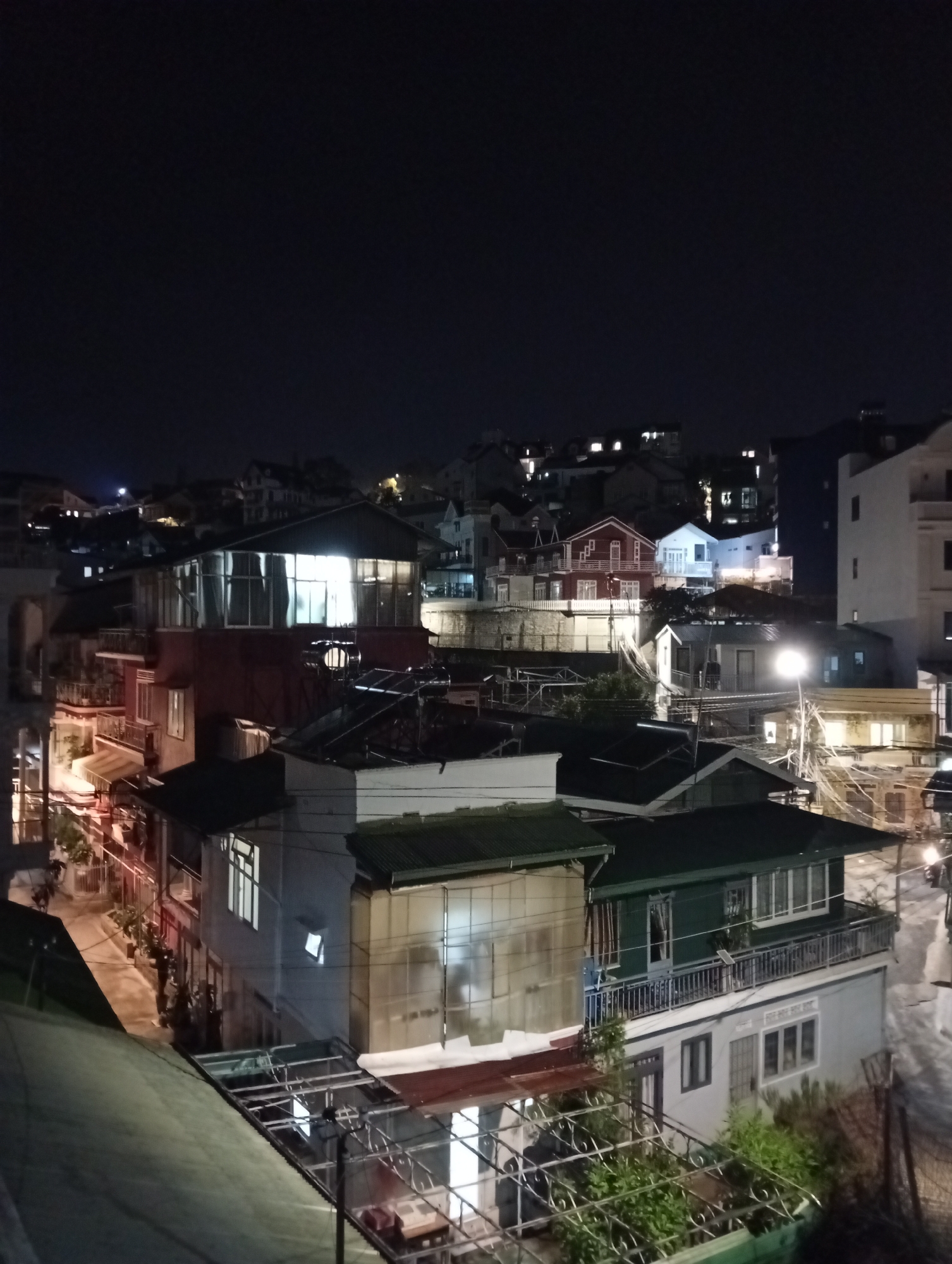 The image depicts a nighttime scene in Da Lat, Vietnam. Numerous buildings with varying heights and architectural styles are arranged on a hillside, illuminated by artificial lights. The sky is dark. A street curves through the buildings, lit by streetlights. Balconies, windows, and roof structures are visible on the buildings. In the foreground, greenery is present near the bottom of the frame. The scene is composed of densely packed buildings.