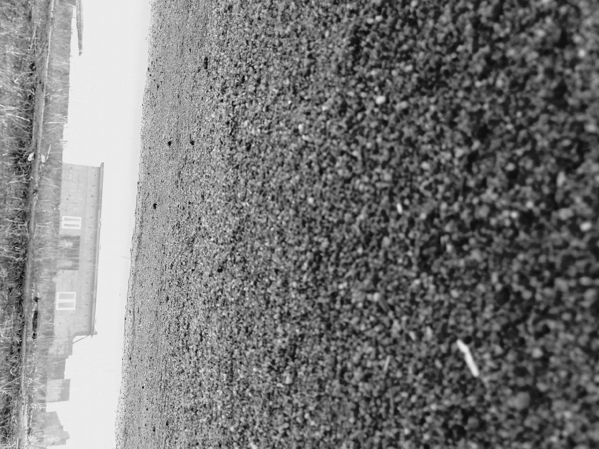 The image shows a black and white, close-up view of a gritty surface, possibly asphalt or small gravel. To the left, there is a building with windows visible. Vegetation, likely grass, is seen to the far left. The location is implied to be in or near Barakin Angundi, Nigeria, based on the prompt.