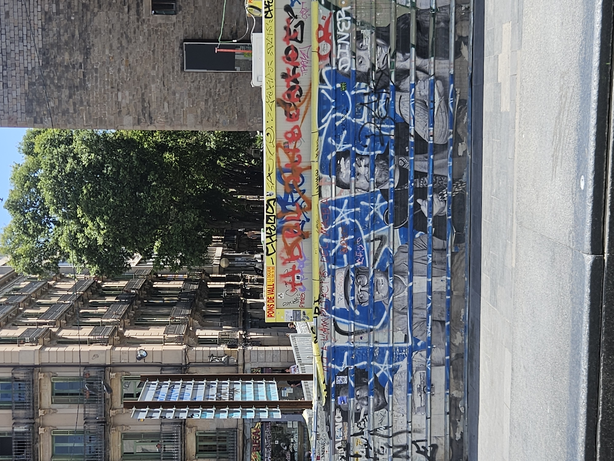 The image captures an urban street scene in Barcelona, dominated by a long barrier or hoarding covered extensively in graffiti. The barrier, running horizontally across the middle of the frame, is composed of vertical panels. The upper section features bright yellow panels covered in red, orange, and black spray-painted tags. On one yellow segment, a sign clearly displays "PONS DE VALL", "LLOGUER" (rental), and a phone number: "93 351 74 08". Below this, a series of blue panels are adorned with white and black graffiti, depicting stylized faces, some wearing glasses, interspersed with abstract designs and text, including what appears to be "DINER".
In the background, a light-colored multi-story building rises, featuring numerous windows, some with green frames, and a distinctive pattern of sun shades or balconies. Further back and to the left, dense green tree foliage partially obscures other structures. To the right and behind the trees, a darker, brick-textured building is visible, with a small window and a black board or sign attached to its facade.
The foreground shows a paved sidewalk or street surface, light grey in color, suggesting a pedestrian area. The scene is illuminated by bright sunlight, casting clear shadows, indicating it is daytime with a clear sky. There is no direct activity taking place, but the vibrant graffiti contributes to a dynamic urban atmosphere.