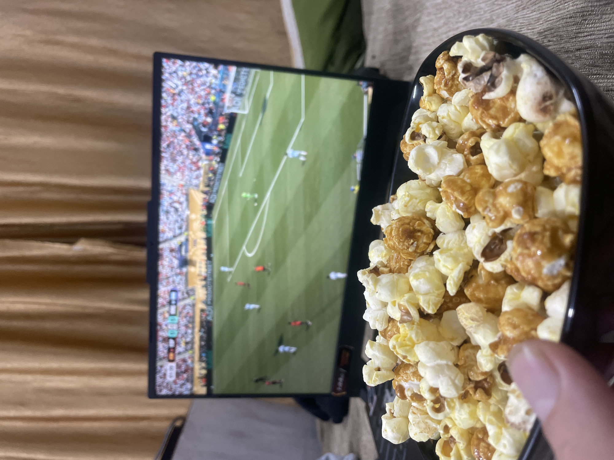 A close-up, high-angle shot features a black bowl filled with a mixture of plain and caramel popcorn in the foreground. A person's thumb is visible on the left side of the bowl, reaching towards the popcorn. In the background, a laptop screen displays a soccer match, with players in white and red uniforms on a green field. The laptop is positioned on a surface that appears to be a bedspread or tablecloth, in front of a backdrop of beige curtains. The overall scene suggests a casual setting, likely an indoor viewing experience of a sporting event, possibly in Bhagsu, India, given the context provided.