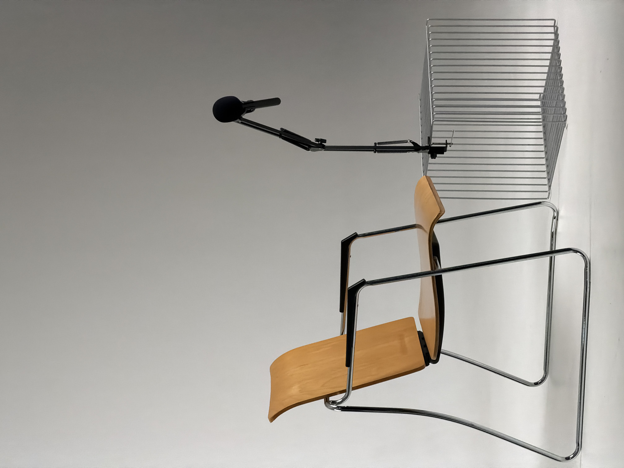 The image shows a minimalist setup with a modern wooden chair with chrome legs and armrests. A black microphone on an adjustable boom arm is attached to a small, cage-like metal table. The background is a plain, neutral gradient, suggesting a studio or controlled environment. There are no people present in the image. The image does not contain any information about the city of Krasnoyarsk, Russia.