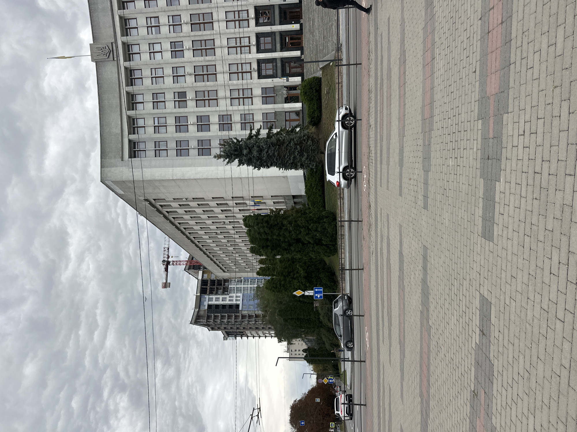 The image depicts a street scene in Ternopil, Ukraine, with a large, imposing concrete building dominating the background. This building, characterized by its repetitive window patterns and a Ukrainian trident emblem atop, appears to be a government or administrative structure. In front of it, a construction site is visible with a red crane. The foreground shows a paved walkway with intricate brickwork patterns. Several cars are on the road, including a silver SUV and a dark-colored sedan, suggesting moderate traffic. The sky is overcast with heavy clouds. A solitary figure, dressed in dark clothing, is walking away from the camera on the sidewalk. The overall impression is one of urban infrastructure and ongoing development under a subdued sky.