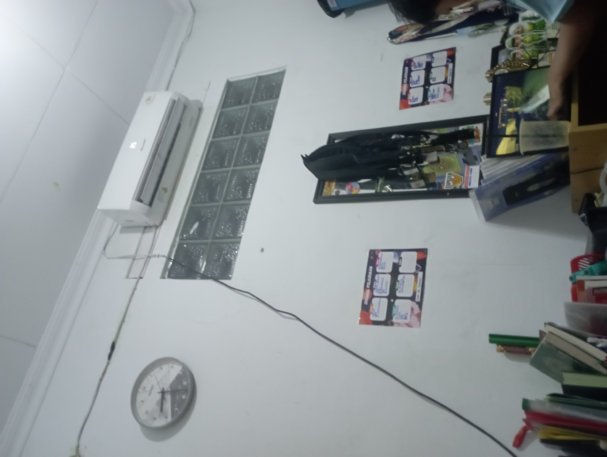 The image depicts an interior scene, possibly a room in Purwakarta, Indonesia, featuring a white wall and a white ceiling. On the wall, a round analog clock with a grey frame is mounted, showing a time around 4:10. Above the clock, a white air conditioning unit is installed. Below the AC unit and to the right of the clock is a small window made of translucent glass blocks.
Several items are placed on a surface in the lower part of the frame. These include stacks of books and papers, a red plastic container, a blue plastic storage bin, and a framed object with a black jacket hanging on it. A calendar with a grid layout is also affixed to the wall. In the lower right corner, a person's arm and shoulder are visible, with their hand appearing to be interacting with a laptop. The laptop screen displays a scene with green elements.
The overall lighting suggests an indoor environment, possibly at dusk or with artificial lighting. The scene appears to be a workspace or a study area.
