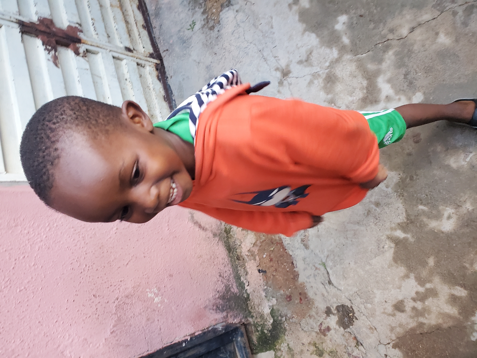 The image shows a young boy, likely of African descent, posing for a photo outdoors in what appears to be a residential area in Jos, Nigeria. The boy is wearing an orange long-sleeved shirt with a black and white striped undershirt and green shorts. He is smiling broadly, with his teeth visible, and looking slightly off-camera.
The setting is a rough, possibly unpaved ground surface with visible cracks and some patches of moss or dampness, suggesting recent rain or a humid environment. To the left of the boy, there is a textured pink wall. Behind him and to his upper left, a section of a white metal fence or gate is visible, with some areas showing signs of rust. The lighting suggests it is daytime, possibly late morning or afternoon, with natural light illuminating the scene. The overall impression is a candid shot of a happy child in an everyday outdoor environment. There is no visible text in the image.