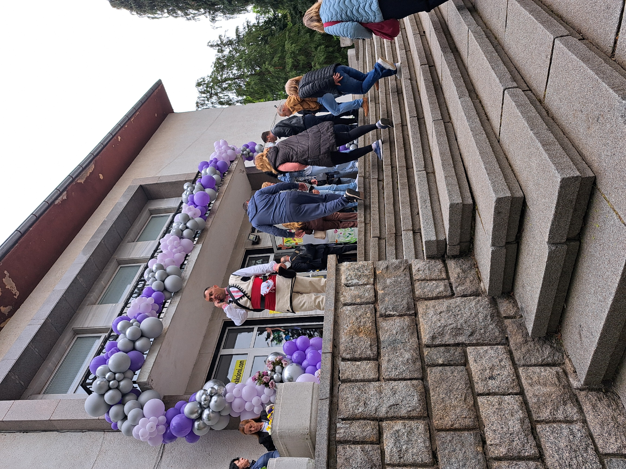 The photograph depicts a building decorated with purple and silver balloons, likely for a celebration or event. A man in traditional attire stands at the entrance, seemingly on a phone call. A line of people is ascending the stone stairs leading up to the building. The location is in the city of Unknown, Unknown.