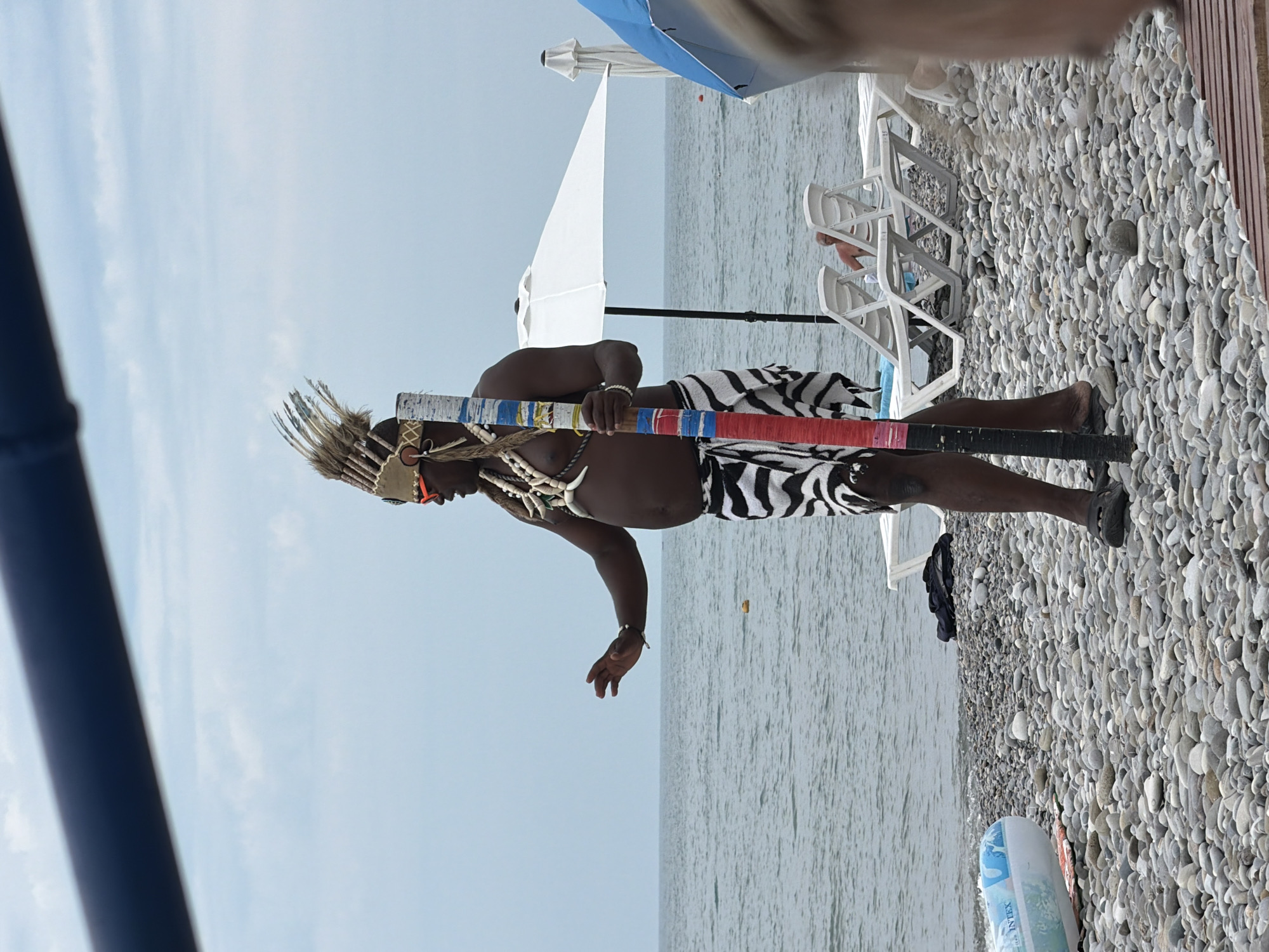 A man in traditional headdress and attire stands on a pebble beach in Sochi, Russia, holding a decorated staff. He wears a headdress adorned with feathers and sunglasses, a necklace made of shells and teeth, and a black and white patterned skirt. The man's right hand is outstretched. Behind him, the Black Sea stretches to the horizon under a cloudy sky. Beach chairs and umbrellas are visible on the sand, suggesting a public recreational area.