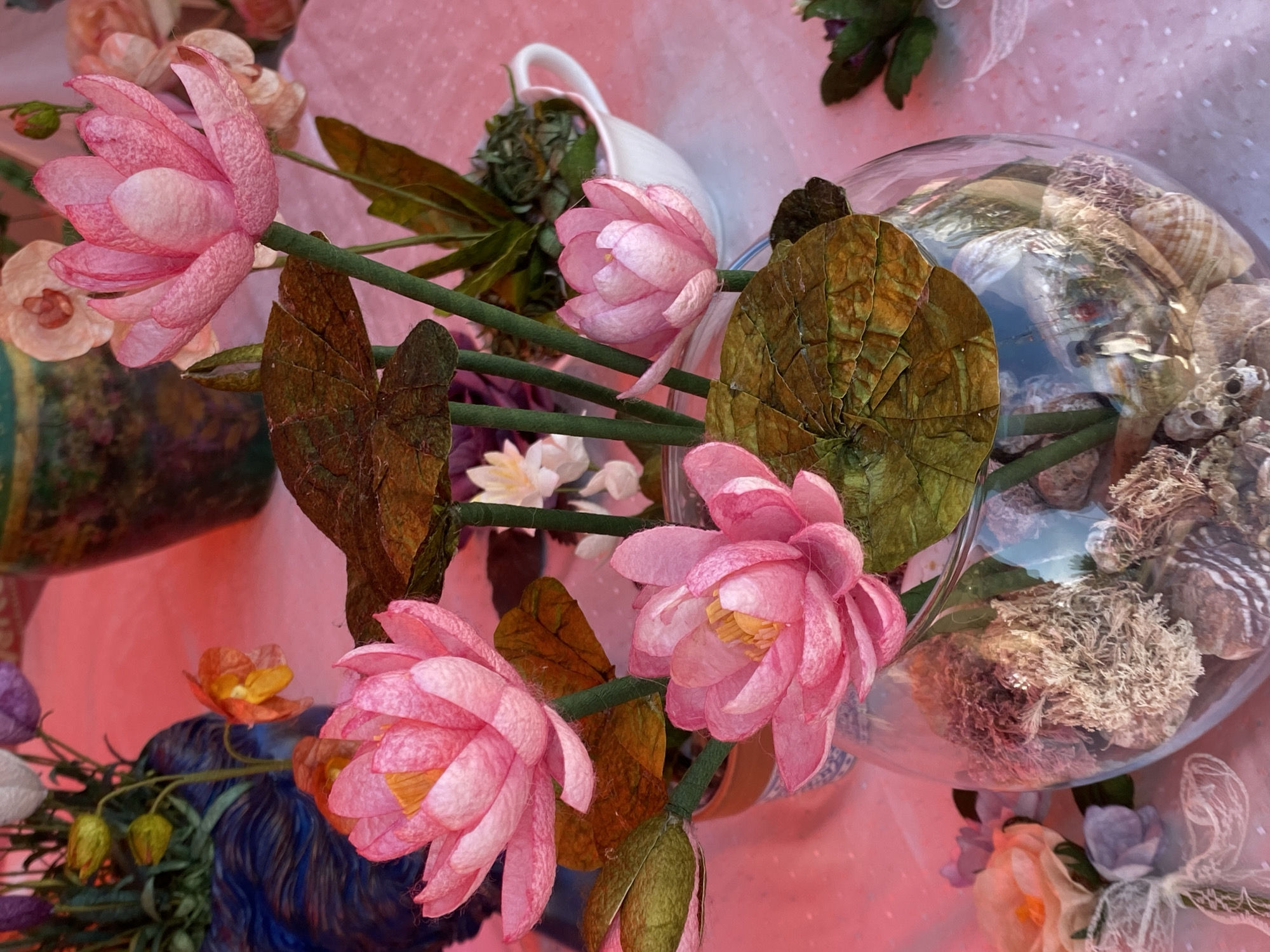 A still life, likely taken indoors, features a variety of floral arrangements and decorative objects. Predominantly, the scene is awash in pink hues due to the lighting and the color of the background. Pink lotus flowers with yellow centers are prominently featured. Their stems and leaves are also visible. Other flowers, in shades of white, orange, and purple, are scattered around, partially obscured. A clear glass bowl filled with stones and what appears to be moss or other natural materials is also visible. There is also a white ceramic pitcher near the back of the arrangement. The location is not evident from the image.