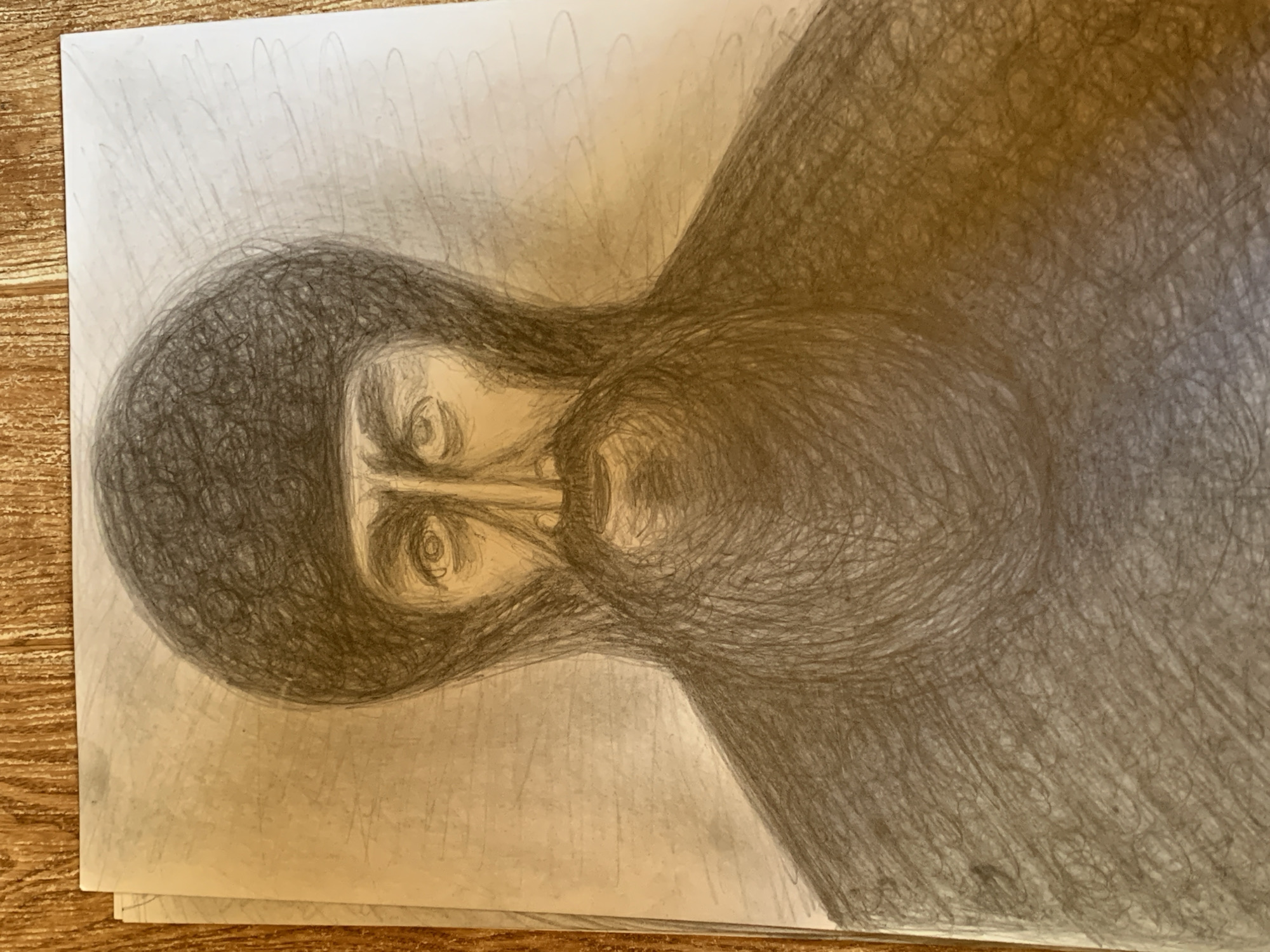 A charcoal sketch on paper depicts a portrait of a man wearing a dark cap and a dark, fur-lined garment. The man has a beard and mustache, and his eyes are open, looking forward. The background of the sketch is a pale, textured surface with faint, scribbled lines. The paper is placed on a wooden surface with a visible grain pattern.