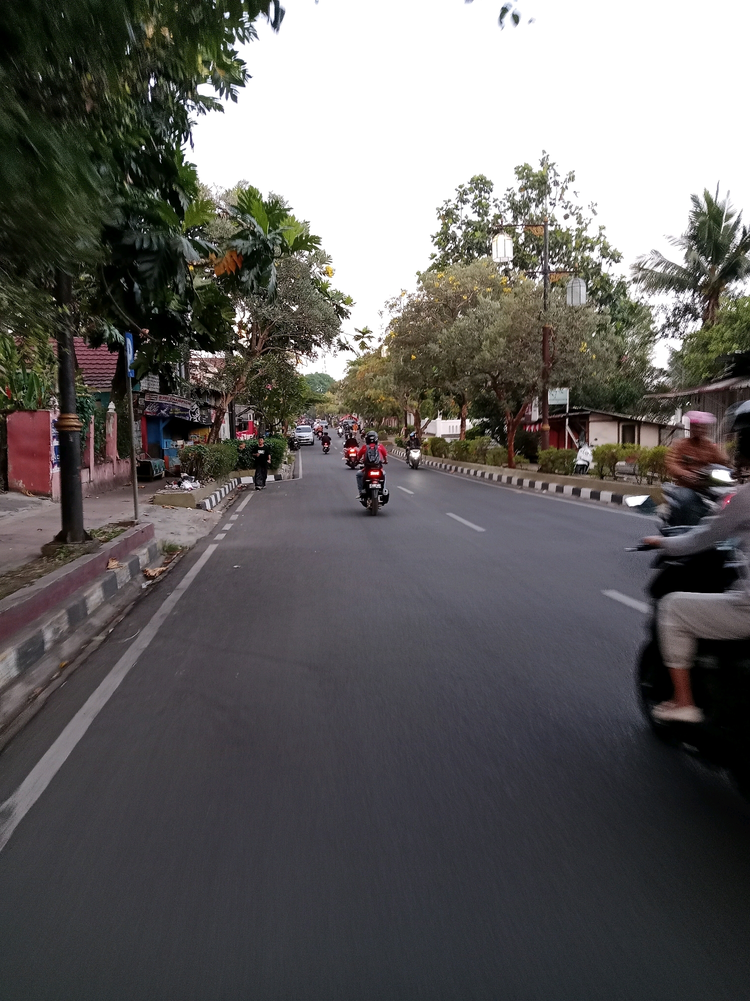 The image depicts a street scene in Cianjur, Indonesia, likely during the day with overcast skies. The perspective is from the side of a moving vehicle, possibly a motorcycle, looking down a paved road. On the left side of the road, a sidewalk is visible with a low retaining wall and trees. Buildings with reddish-brown roofs and some businesses line the sidewalk. On the right side of the road, there are trees and more buildings. The road itself is busy with traffic, primarily motorcycles, some with riders wearing helmets. A few cars are also present in the distance. The overall impression is of an active urban environment.