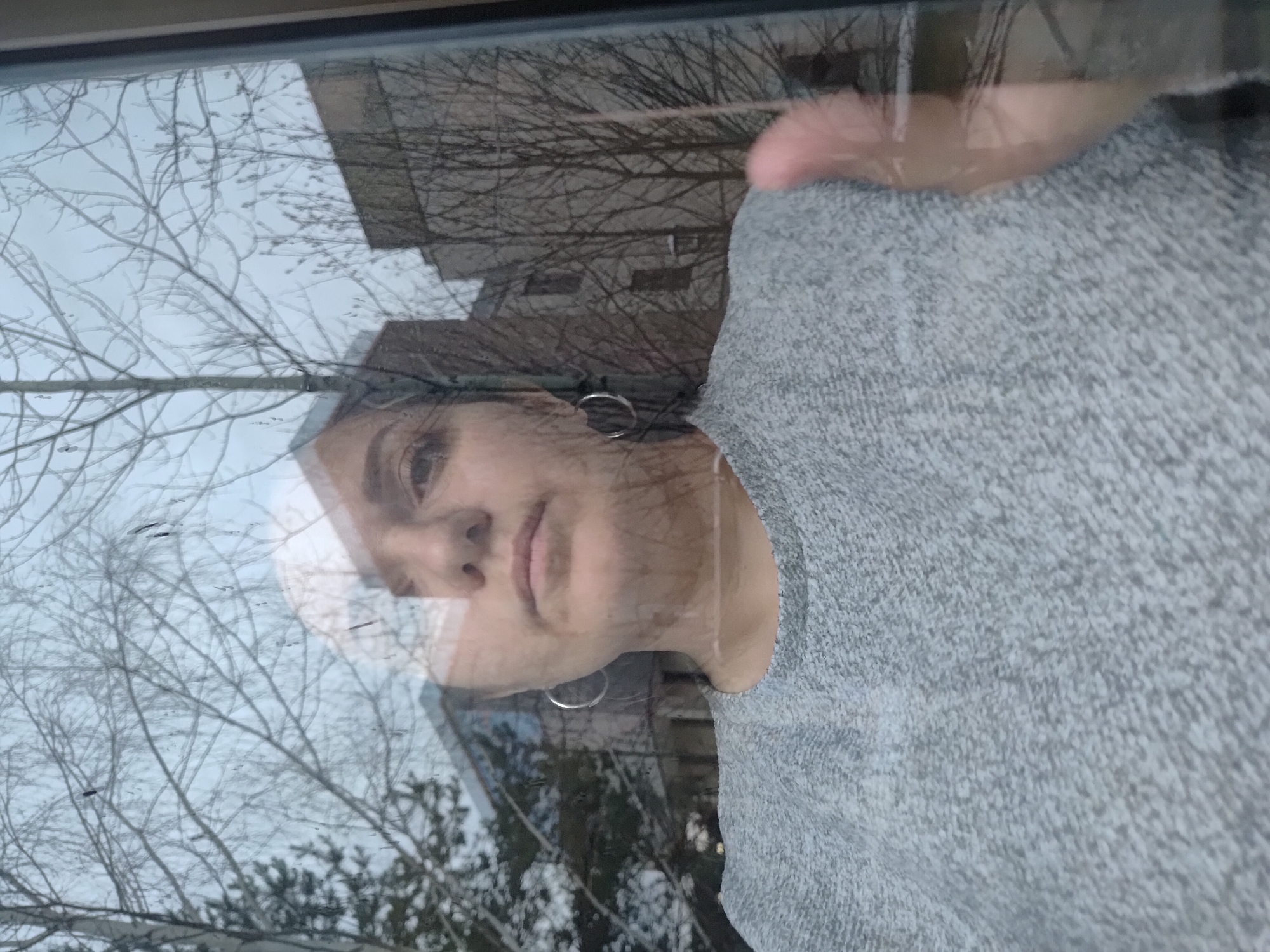 The image, rotated 90 degrees clockwise, captures a reflection of a person in a window, superimposed over an outdoor scene. On the right side of the frame, the upper body and face of a woman are visible. She has dark hair and a neutral to slightly contemplative expression, with her gaze directed slightly away. She wears a textured grey sweater or hoodie, and a silver hoop earring is visible on her left ear.
Reflected in the glass, an outdoor view serves as the background for the woman's reflection. This scene is dominated by numerous bare tree branches, intertwining against a light, overcast sky. In the deeper reflection, indistinct structures or buildings can be discerned among the trees. The absence of leaves on the trees indicates a colder season, likely late autumn, winter, or early spring. The lighting is soft and diffused, characteristic of an overcast day.
No specific activity is overtly depicted beyond the woman's presence near a window, possibly observing the exterior or interacting with her reflection. There is no visible text in the image. The scene, with its bare trees and subdued light, is consistent with the climate of Markovskii, Russia during colder seasons.
