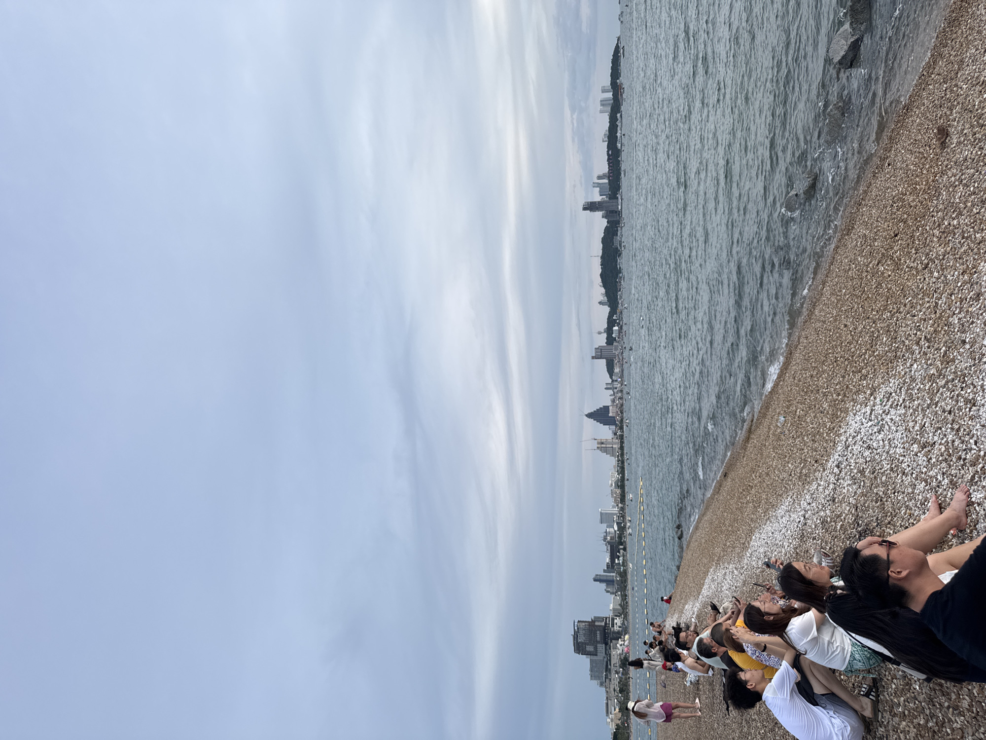 A group of people are sitting on a pebble beach in Pattaya City, Thailand, facing the ocean. In the background, a cityscape with numerous buildings, including high-rises and cranes, is visible across the water. The sky is overcast with clouds. The sea is moderately choppy with some waves reaching the shore. Some boats are scattered in the water. The people are dressed in casual clothing, and some are looking at their phones. The scene suggests a relaxed, possibly evening, atmosphere.