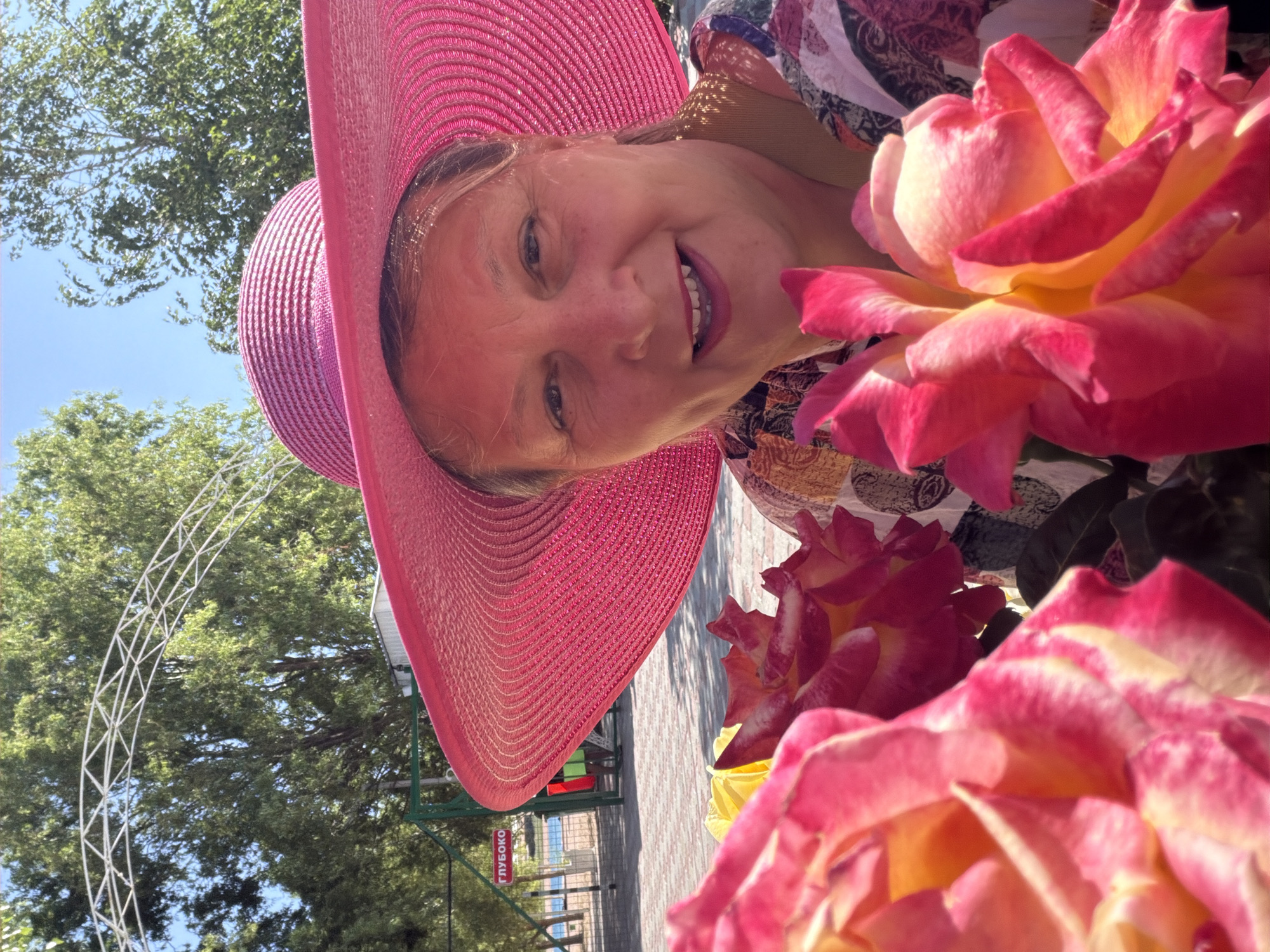 A middle-aged Caucasian woman is posing for a close-up selfie in an outdoor environment. She is wearing a wide-brimmed pink straw hat that obscures the top portion of her head and frames her face. Her eyes are looking directly at the camera, and she has a smile with her mouth open, revealing her teeth. She is wearing a colorful, patterned blouse. In the foreground, large pink and yellow roses are partially out of focus, creating a vibrant and somewhat abstract framing element. Behind her, trees with green foliage are visible against a bright blue sky. To the left, a white, arched metal structure is visible, possibly part of a garden or park. Further in the background, a green fence encloses an area with a sign in Cyrillic script that reads "ГЛУБОКО" (meaning "Deep"). Beyond the fence, there appears to be a body of water, possibly the sea, and a few people are standing near the fence. The overall impression is a cheerful portrait taken in a sunny, garden-like setting.