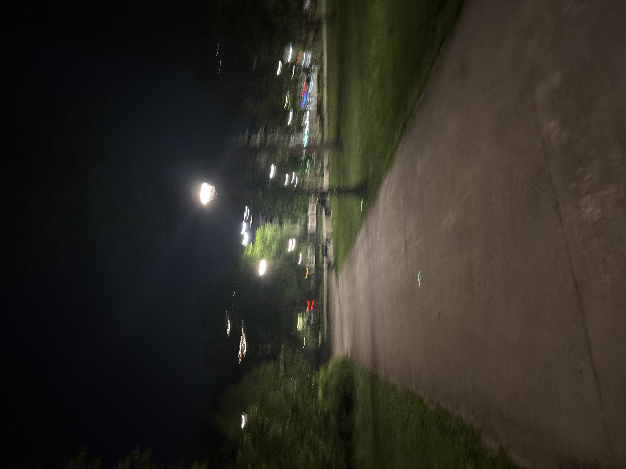 A nighttime scene depicts a paved pathway flanked by grassy areas and trees. The pathway, illuminated by streetlights, extends into the distance. Distant lights, some blurred, are visible among the trees. The left side of the frame is dominated by darkness. The scene is likely taken in a park or green space within a city at night. The location is Perm, Russia.