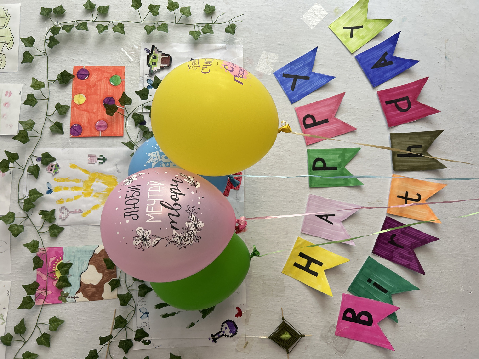 The image shows a close-up of a wall decorated for a birthday celebration. There are several colorful drawings and artwork by children pinned to the wall, including a handprint and drawings of nature scenes and abstract shapes. Artificial ivy vines are draped across the top of the wall and around the decorations. In the foreground, a collection of balloons in pink, green, blue, and yellow are hanging, with the pink balloon featuring text in Cyrillic script and floral motifs. Below the balloons, a "HAPPY BIRTHDAY" banner is strung across the wall, made of individual pennant flags in various bright colors, each bearing a letter. The overall scene suggests a festive and personalized celebration, likely in a child's room or a classroom setting. There is no indication of the specific location within the city of Kerch, Ukraine.