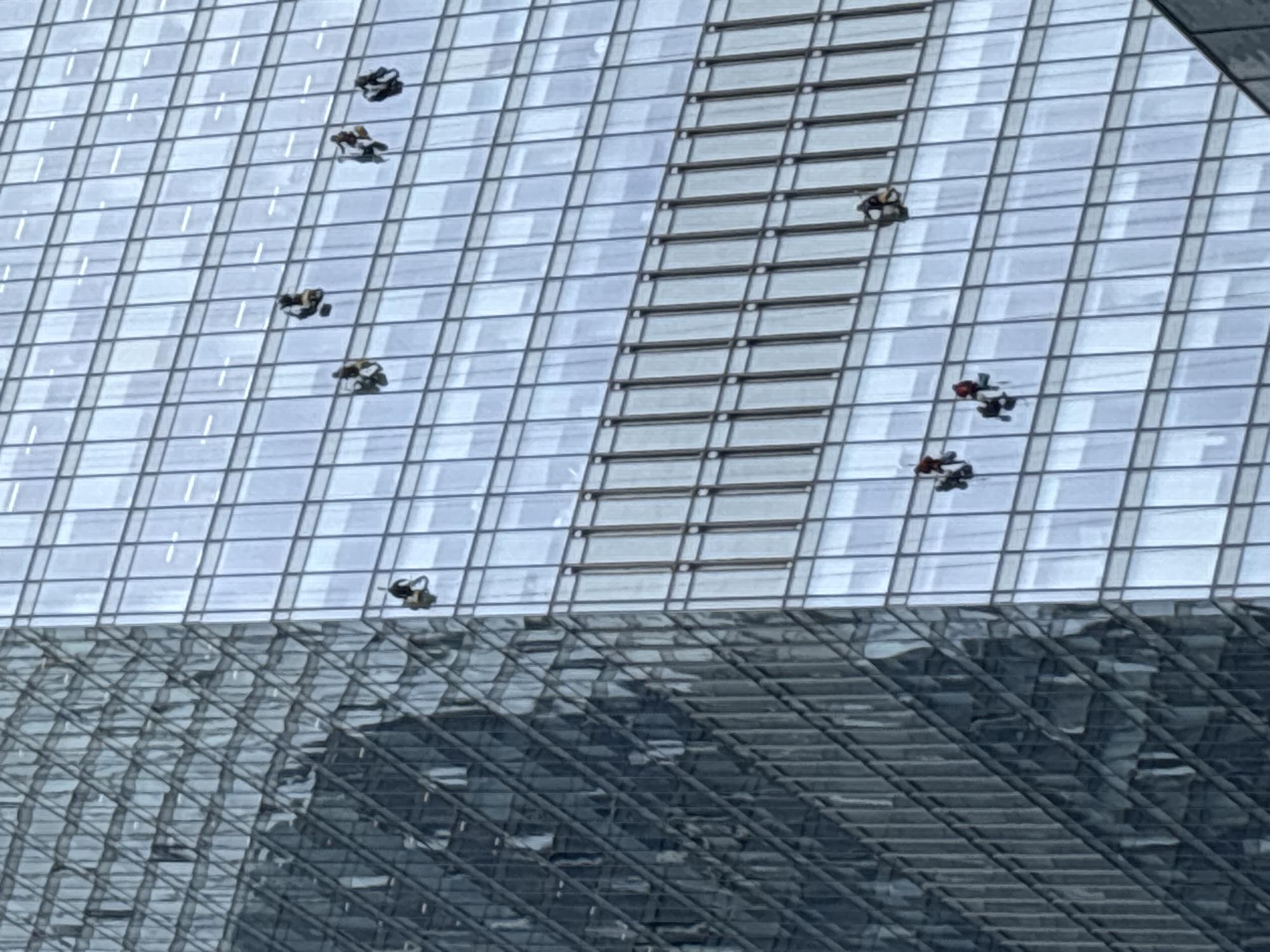 The image shows a partial view of a skyscraper in Moscow, Russia, featuring a large glass facade with visible grid-like window patterns. Several window washers are suspended on ropes, working on cleaning the building's exterior. The reflection of the surrounding cityscape is visible on the lower section of the building.