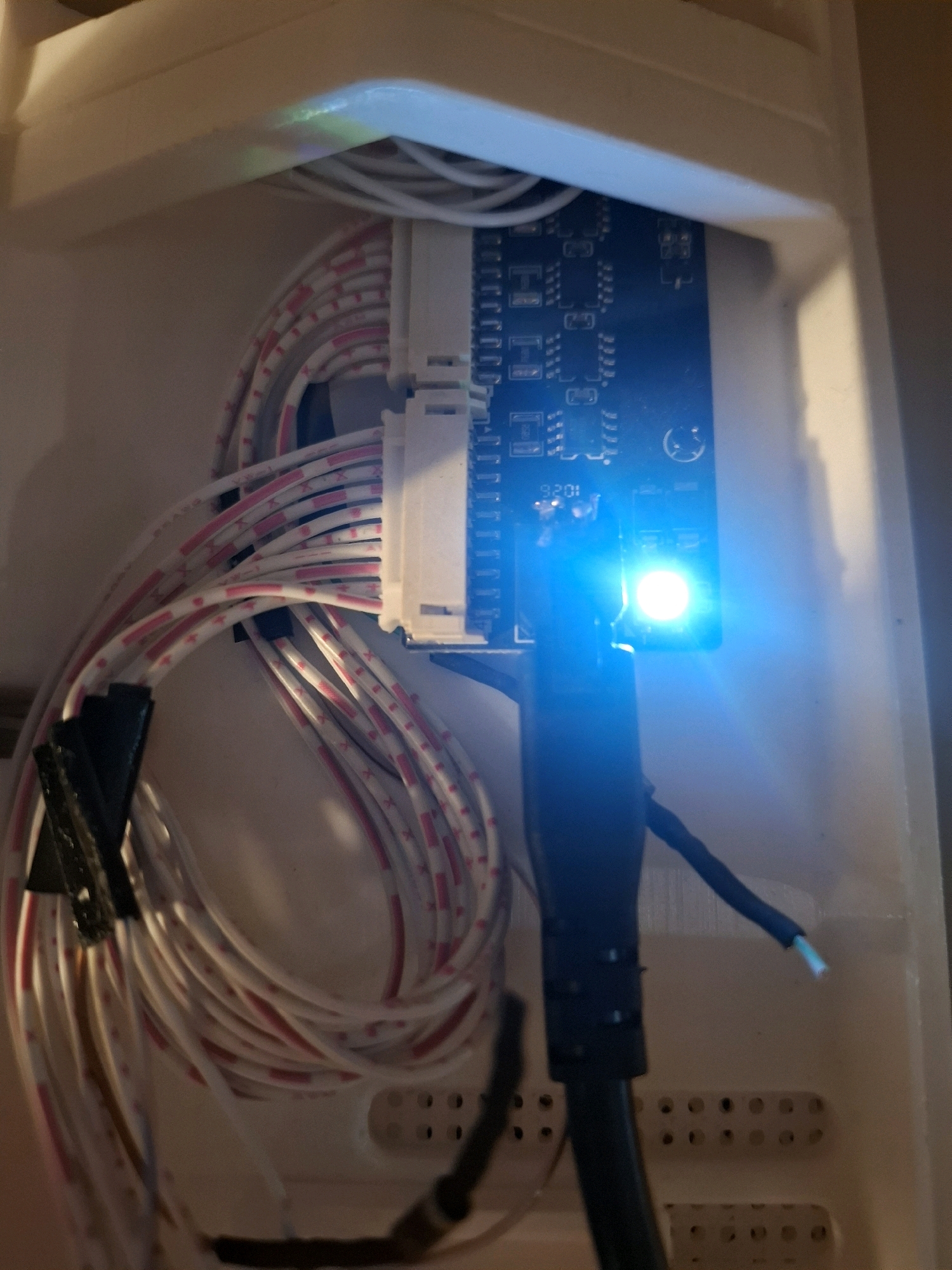 The image shows a close-up view of electronic components inside a white plastic enclosure. A blue circuit board is visible, with a bright blue LED illuminated on its surface. A bundle of white wires with pink stripes is connected to the circuit board. The enclosure appears to be part of a larger device. There are no people or any indications of a specific location in the city of Tyumen, Russia. The scene is static and shows electronic hardware.
