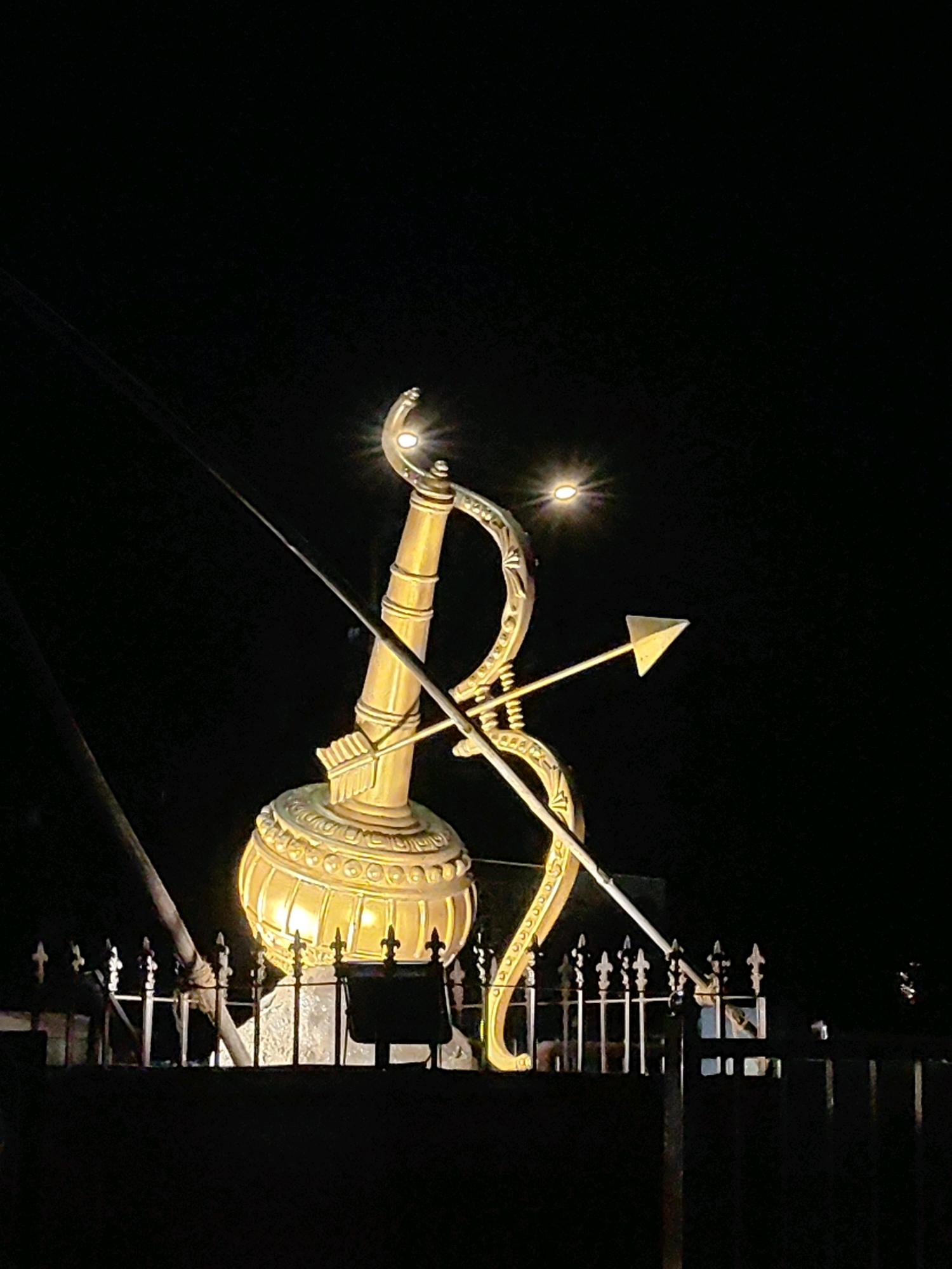 This is a nighttime, medium-shot, eye-level photograph of a large golden statue depicting a bow and arrow, likely representing a deity or mythological figure, illuminated by spotlights. The statue is positioned in front of a dark background, presumably the night sky. A fence is visible in the foreground, with some parts of the statue's base appearing to rest on or behind it. The statue itself is intricately detailed, with a large, bulbous base and a curved bow that extends to the right. An arrow is shown horizontally, passing through the bow. Two bright lights are visible on the statue, seemingly part of its illumination system. The overall impression is one of grandeur and symbolism, illuminated against the darkness.
While the image does not directly provide information about its specific location within Mumbai, India, the visual elements such as the style of the statue and the fence could potentially be linked to religious sites or cultural landmarks in the city. Without further context or distinguishing features in the background, pinpointing the exact location is not possible from the image alone.