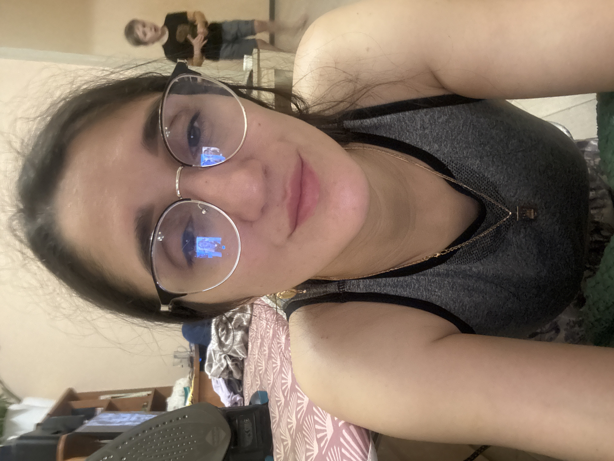 A selfie shot shows a woman with fair skin, wearing round-frame glasses and a gray tank top with black trim. She is wearing a gold chain necklace. Her dark hair is pulled back. The reflection in her glasses suggests she is looking at a screen.
In the blurred background, a person in a black t-shirt and gray shorts stands near a light-colored wall. An ironing board and iron are visible in the foreground. Based on available information, the location cannot be determined to be Tobolsk, Russia.