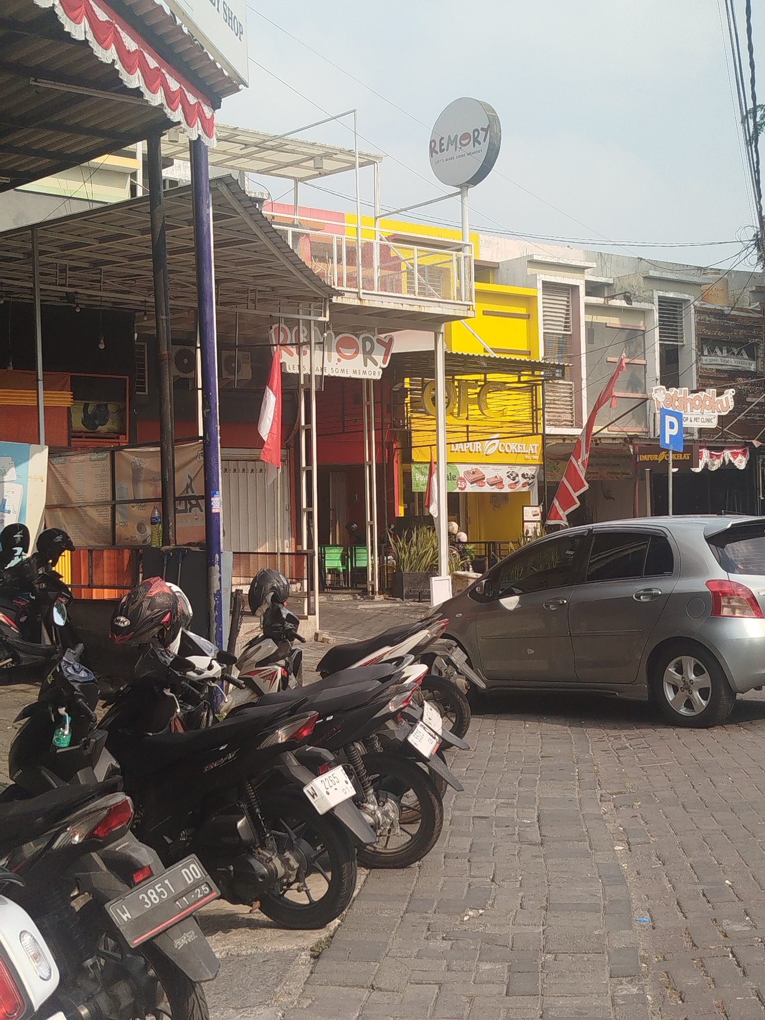 The image depicts a street scene in Yosowilangun, Indonesia, featuring several parked motorcycles and a silver hatchback car. The foreground is dominated by a row of motorcycles, with their license plates visible, indicating that at least two are registered in the W series. One motorcycle's license plate reads "W 3851 DQ" and another "W 2265". Behind the motorcycles, a silver car is parked, partially obstructing the view of the storefronts.
The background reveals buildings with various architectural styles and signage. One building has a prominent yellow facade with the words "DAPUR COKELAT" (Chocolate Kitchen) visible on a sign, along with images of chocolate products. Another sign features a circular logo with the word "REMORY" and the tagline "LET'S MAKE SOME MEMORY." An Indonesian flag, red and white, is also displayed. A sign for "Petshopku" with a parking symbol is present. The street is paved with cobblestones. The sky is overcast, and the lighting suggests it is daytime. No people are clearly visible interacting within the scene, though some figures may be obscured within the buildings or on motorcycles.