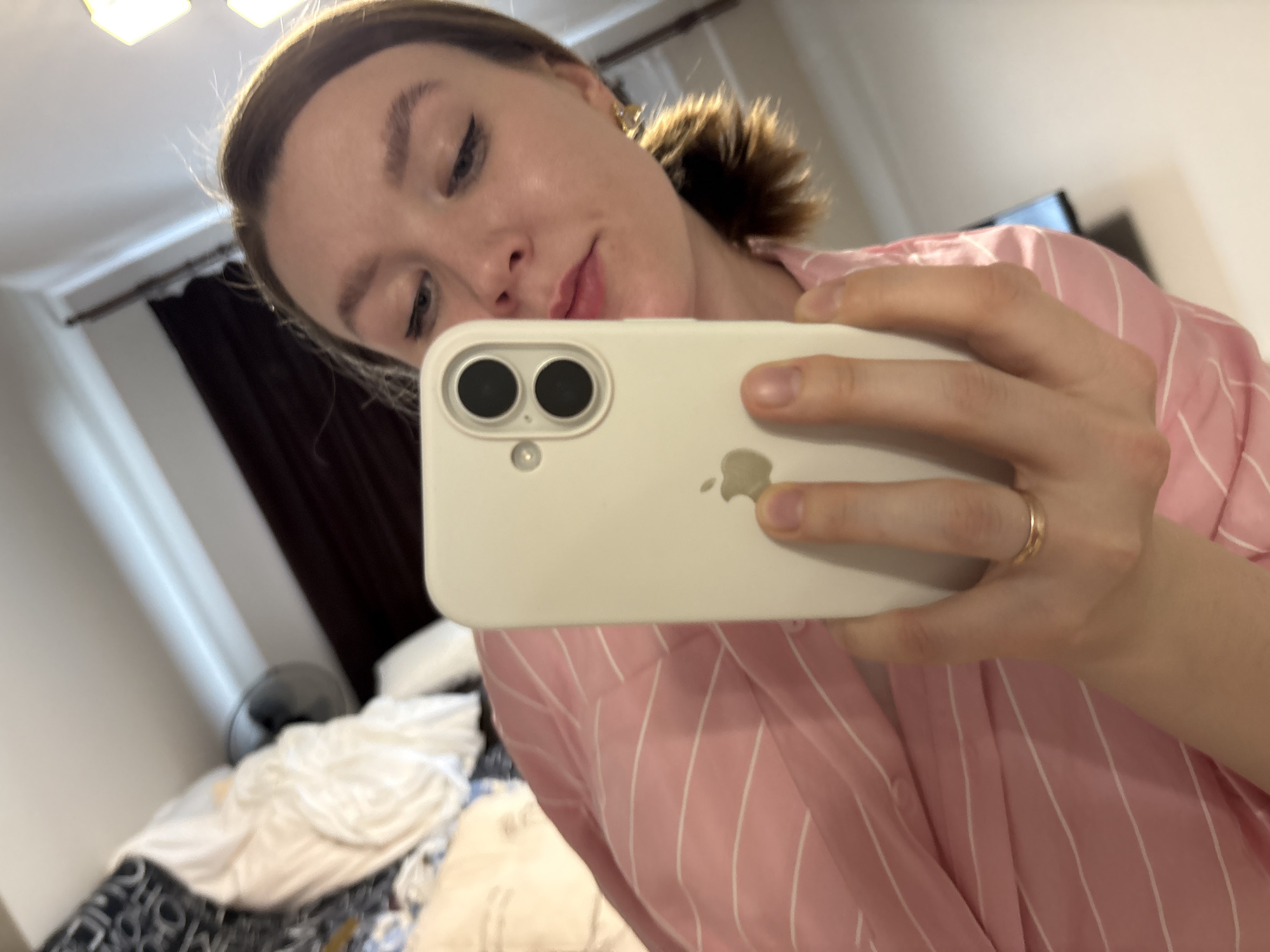 A woman is taking a selfie indoors, likely in a bedroom. She's wearing a pink and white striped pajama top. She's holding a white iPhone in front of her face. A messy bed is visible in the background along with a black fan. The location is impossible to determine based on the provided information.