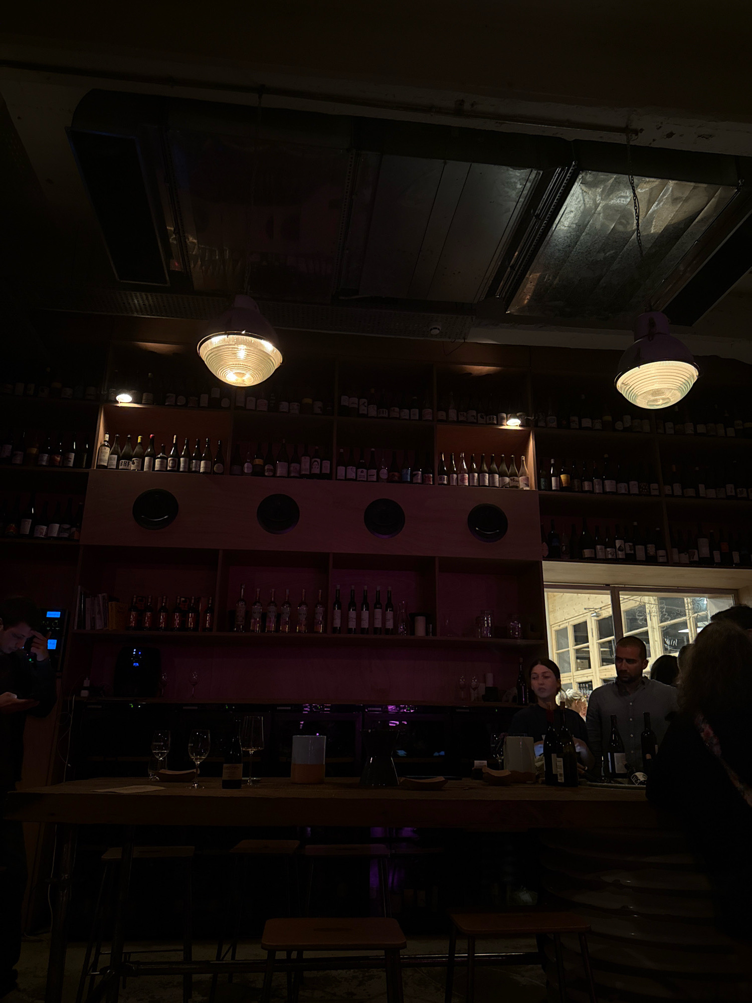 A dimly lit interior of what appears to be a wine bar or restaurant in Tbilisi, Georgia, is captured in this photograph. In the foreground, a wooden bar top is visible, with several wine glasses and bottles sparsely placed on it. Three stools are tucked beneath the bar, suggesting a seating area. A man in dark clothing is standing to the left of the frame, looking at a device in his hand. Behind the bar, a woman is standing and appears to be interacting with a patron or preparing drinks. To her right, a man in a light blue shirt is also visible. The background is dominated by shelves filled with numerous bottles, likely wine, organized in a systematic manner. Two large industrial-style pendant lights hang from the ceiling, illuminating the space with a warm glow. The overall ambiance is one of relaxed socializing, consistent with a bar or restaurant setting.