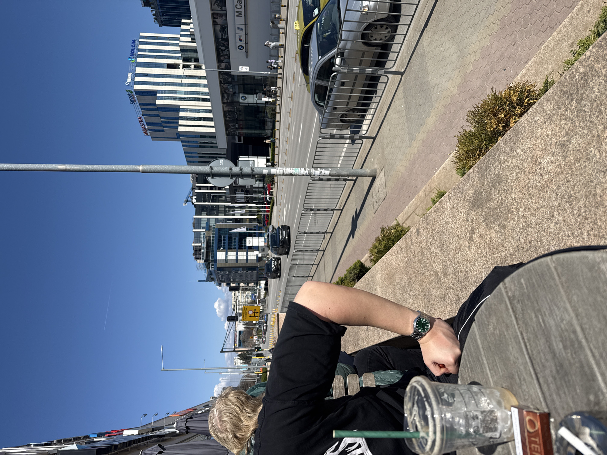 A medium shot, taken in Sofia, Bulgaria, depicts a person lounging on a wooden bench in an urban environment. The individual, seen from an elevated vantage point, is wearing a black t-shirt and pants, and a green-faced watch is visible on their wrist. An empty Starbucks cup with a green straw, a box of Tic Tacs, and an object are positioned on the table in front of the person.
In the background, a busy street runs parallel to the bench, with cars and pedestrians visible. Modern buildings, including one with the Holcim sign and the BMW car center, line the street, indicating a commercial area. The sky above is clear and blue, indicating a sunny day.