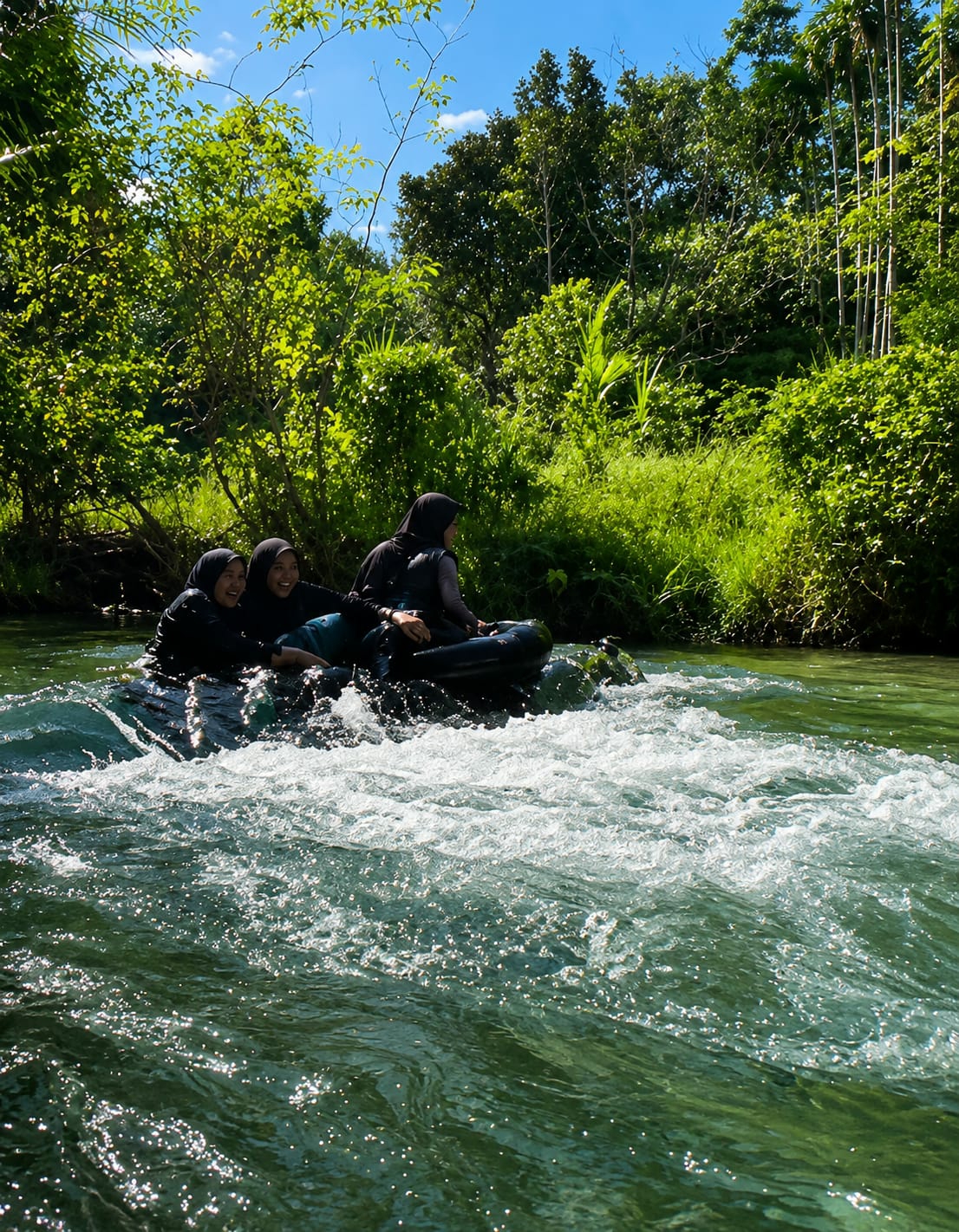 Four individuals, three female and one male, are riding in an inflatable raft on a river. The raft is dark-colored and appears to be moving through choppy water. The three female individuals are wearing dark hijabs and dark clothing. The male individual is to the right of the raft. The raft is positioned in the foreground of the image, with the river flowing rapidly around it. The background consists of lush green vegetation and trees, with a clear blue sky visible above. The scene appears to be set in a natural, tropical environment. This location is identified as Silaiyajae, Indonesia.