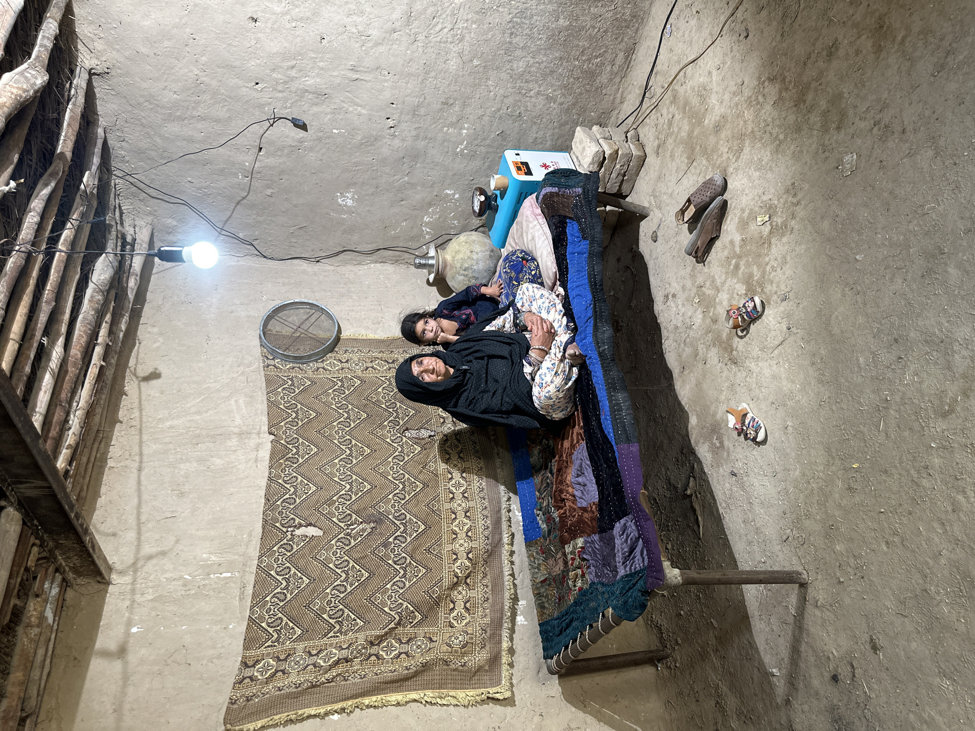 The image depicts an indoor scene in what appears to be a simple dwelling in Quetta City Tehsil, Pakistan. An elderly woman wearing a dark head covering and patterned clothing is lying on a bed alongside a young child. The bed is adorned with colorful patchwork and placed against a wall covered with a traditional patterned rug. Various objects are scattered around the room, including shoes, a water cooler, and a large container. The room is dimly lit by a single light bulb, and the walls appear to be made of mud or clay.