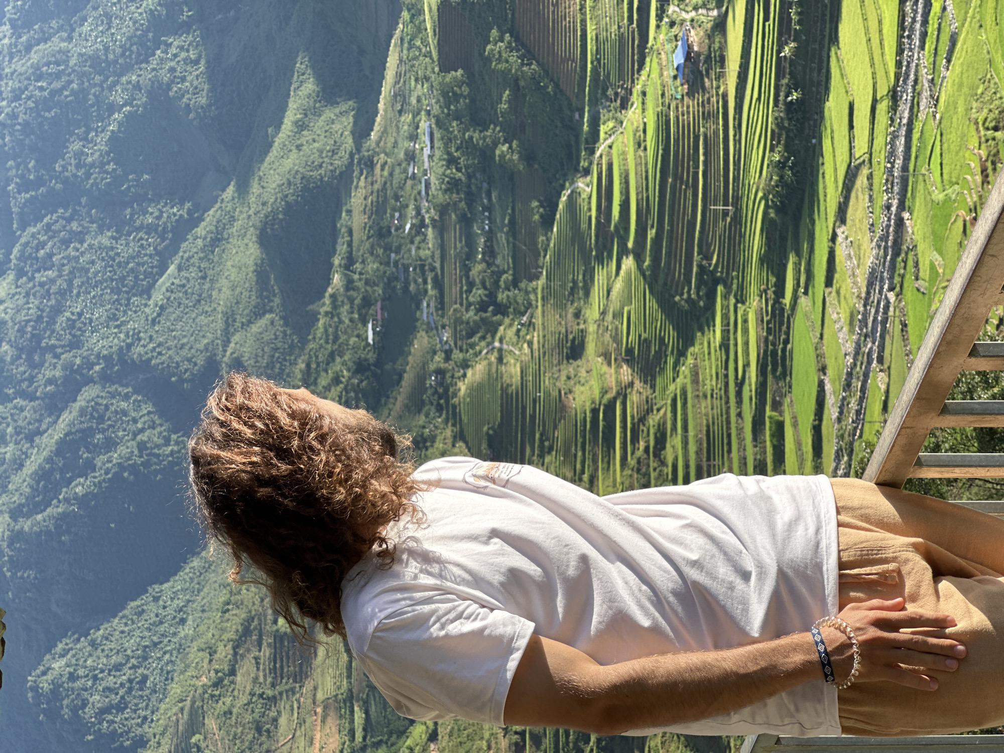 A person with long, curly brown hair, wearing a white t-shirt and light brown pants, stands on a balcony overlooking a mountainous landscape in Tú Lệ, Vietnam. The individual is positioned with their left side slightly towards the viewer, looking towards the right, observing the extensive green rice terraces carved into the hillsides. Two bracelets, one blue and white patterned, and one beaded (gold/pearl-like), adorn their left wrist. In the background, numerous vibrant green rice paddies and terraced fields descend into a valley, interspersed with small houses featuring blue or brown roofs. Beyond the immediate cultivated areas, lush, forest-covered mountains rise steeply, appearing hazy in the distance under bright natural sunlight. A wooden railing is visible in the lower right foreground, indicating an elevated viewing point.