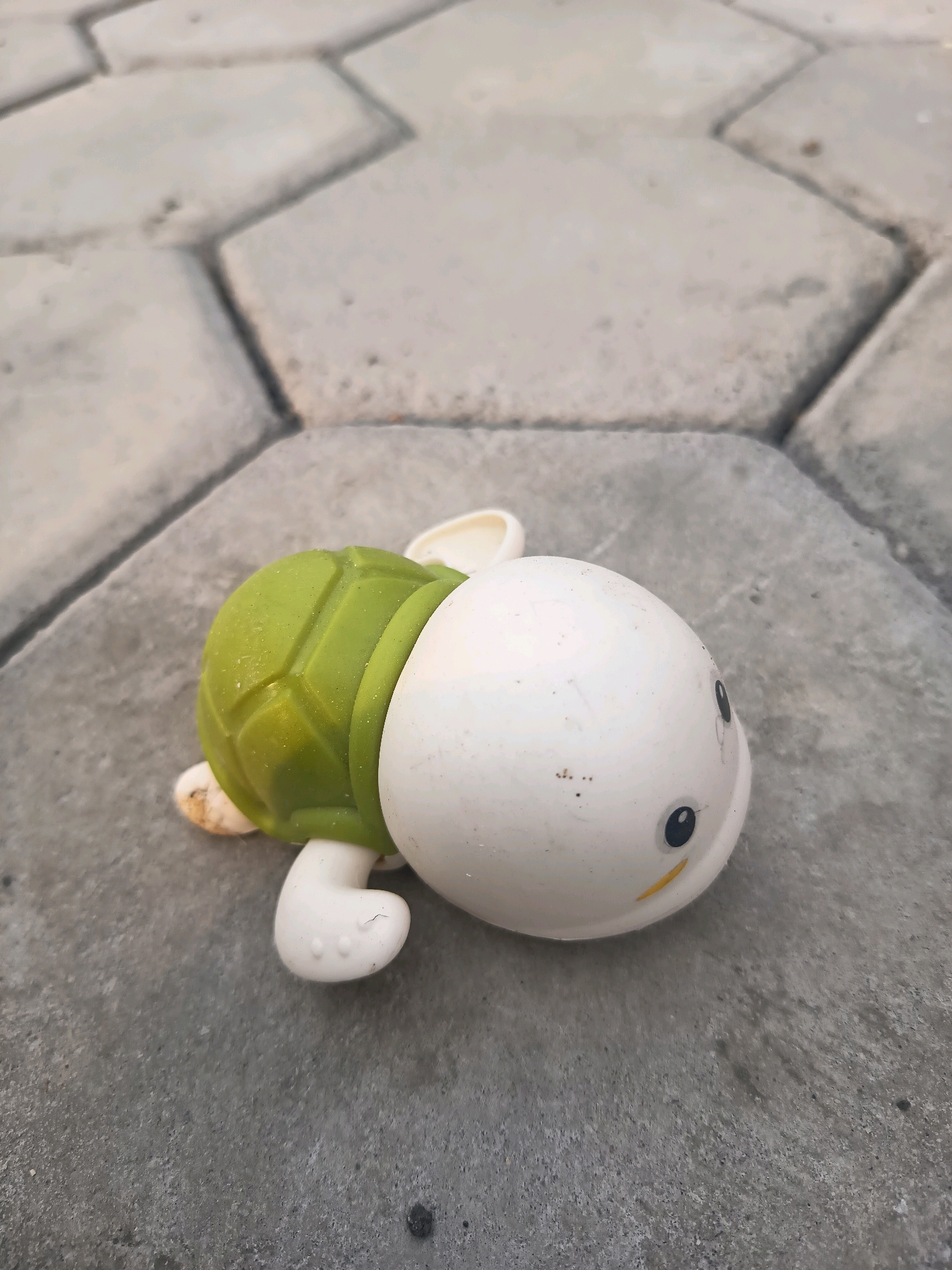 A white and green rubber turtle toy lies on a concrete paver surface. The turtle is facing to the right, with its head turned slightly towards the viewer. It has a white head with black eyes and a yellow smile. The turtle's shell is green and textured. The concrete pavers are light gray and arranged in a hexagonal pattern. The background is out of focus. There is no indication of location in the city of Sidorejo, Indonesia.