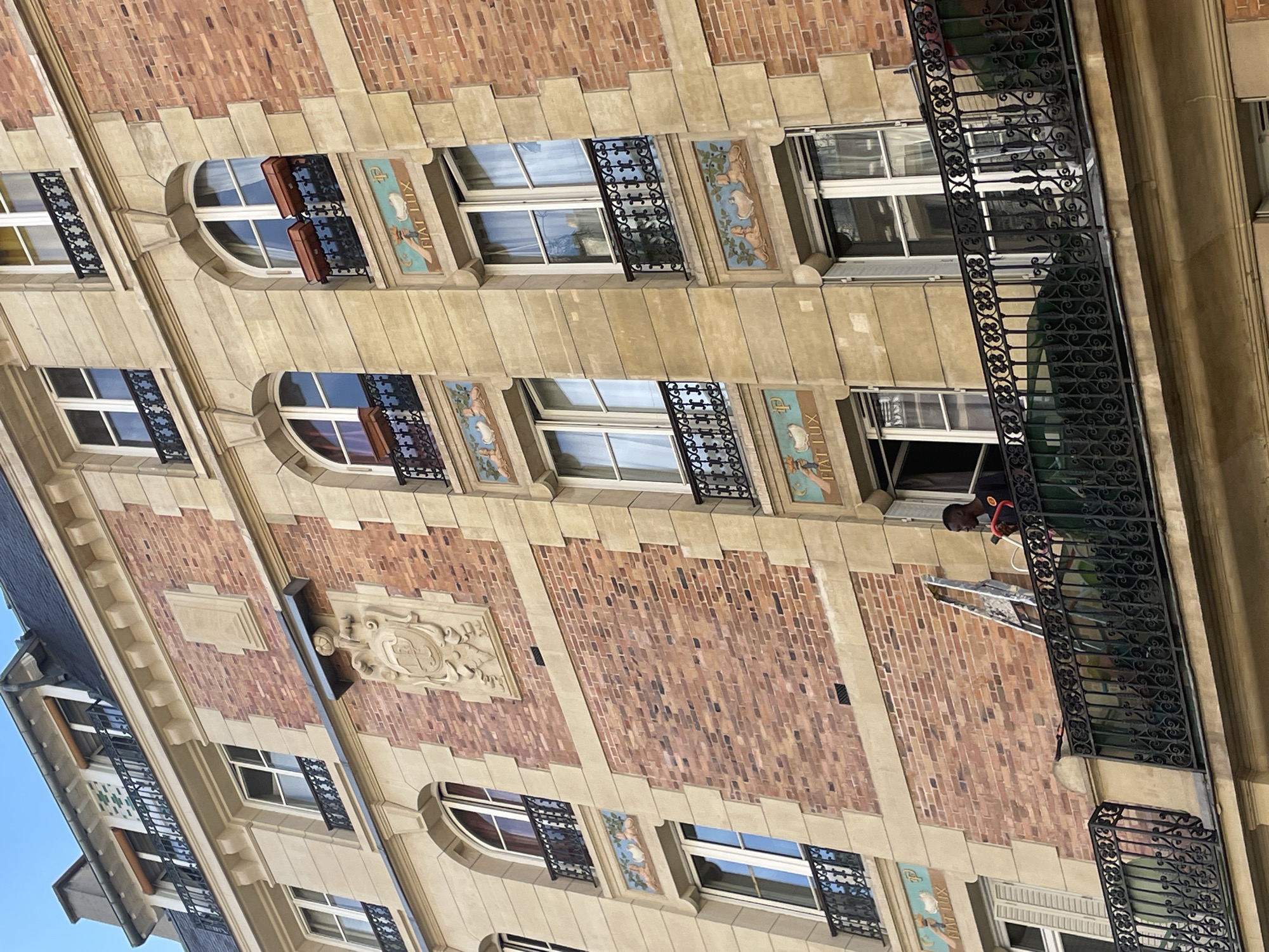 The image displays the facade of a multi-story building, characteristic of Parisian architecture, captured during daylight hours under a clear blue sky. The building features alternating sections of light-colored stone trim and reddish-brown brickwork.
The facade is punctuated by numerous windows, some rectangular and others with arched tops, all framed in white. Many windows are adorned with small, intricate wrought-iron balconettes. Between these windows, decorative ceramic or painted panels depict classical figures or motifs in shades of blue, green, and brown. The Latin phrase "FIAT LUX" (Let there be light) is clearly visible on several of these panels, along with a stylized letter "P." Two windows on an upper level are fitted with brown flower boxes.
On one of the central floors, a man is visible on a larger, continuous balcony with an ornate wrought-iron railing. He is wearing a dark shirt and an orange safety harness, suggesting he is performing maintenance or cleaning work on the building's exterior. Below him, some equipment, possibly a ladder or platform, is partially visible. Further down the facade, a decorative stone crest or emblem is set into a section of brickwork.