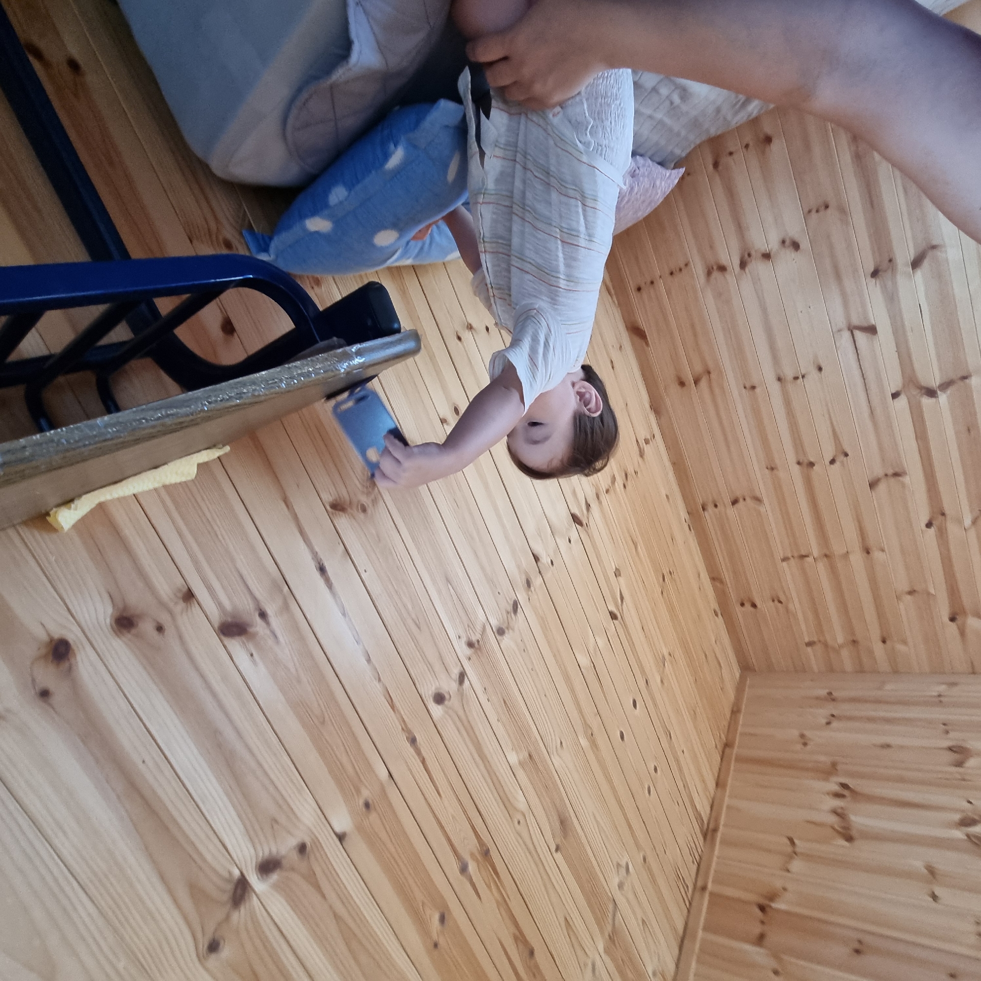 The image shows a young child playing with a blue phone case on a wooden floor. The child is wearing a white shirt with thin stripes. A person's arm is visible in the upper right of the frame, suggesting they are holding the child or the camera. In the background, there are pillows and what appears to be a bed. The scene is indoors, likely in a home, with walls and flooring made of light-colored wood. The lighting suggests it could be daytime. The child appears engaged with the phone case. There is no visible text in the image.