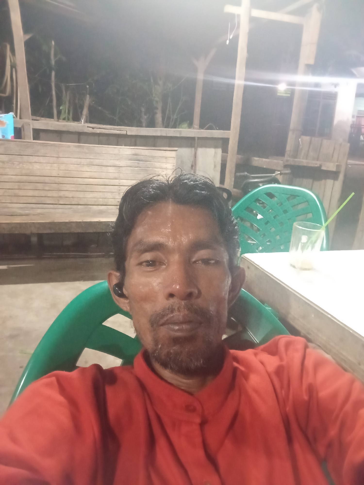 A man with dark hair and a beard is pictured sitting in a green plastic chair in what appears to be an outdoor, dimly lit setting, possibly a warung or simple eatery. He is wearing a red collared shirt and has a black earbud in his left ear. Behind him, there is a wooden bench and more green chairs. A white table is to his right, with a glass containing a beverage and a green straw on it. The background shows wooden structures and vegetation, suggesting a rural or semi-rural environment. The image quality is somewhat grainy, indicating it might have been taken with a mobile phone at night or in low light. The location is Lhokseumawe, Indonesia.