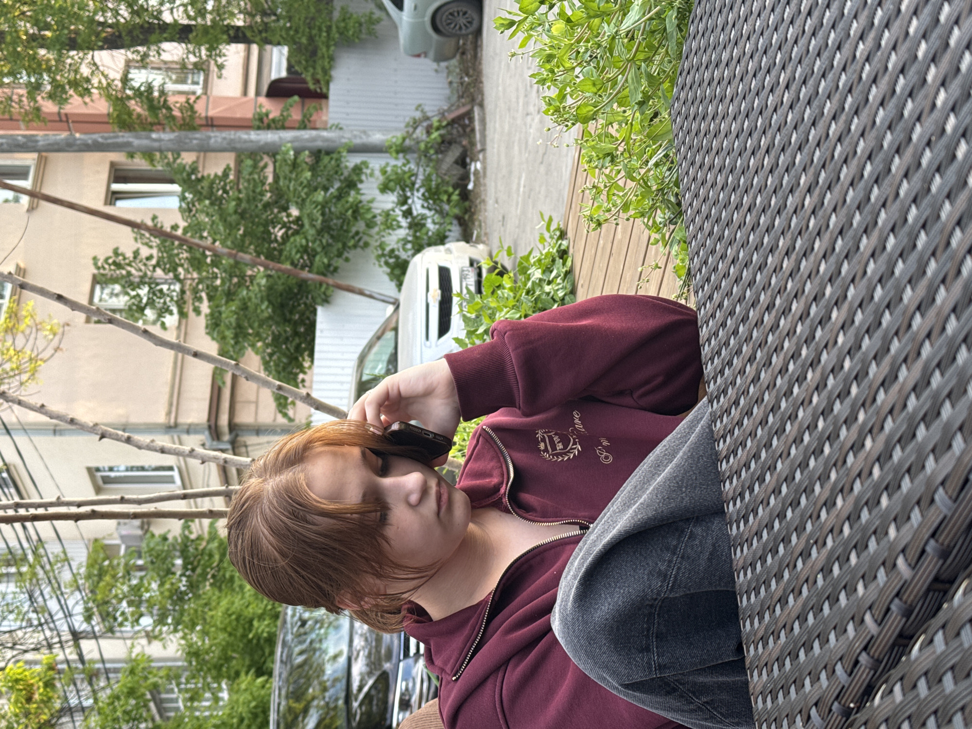 A young person with reddish-brown hair is captured in a profile view, seated outdoors, and engaged in a phone call. They are wearing a maroon zippered hoodie with dark gray jeans. Their right hand holds a dark smartphone to their ear, and their gaze is directed downwards, suggesting a thoughtful or focused demeanor during the call.
The setting appears to be an outdoor patio or balcony, indicated by the dark brown woven wicker-like surface of a table or chair armrest in the foreground. The background reveals an urban or residential environment with multi-story buildings, some with visible windows, and various green trees and bushes. Bare tree branches are prominent in the midground. Parked vehicles, including a white van and another light-colored car, are visible on what seems to be a street or paved area.
The light is diffused, suggesting it is daytime, possibly a cloudy day or late afternoon. On the left chest of the maroon hoodie, there is an embroidered crest. Although somewhat indistinct, the crest contains interwoven letters that appear to be "P. D." or "D. D.", with script text underneath that could read "Clare" or "College." The overall scene suggests a quiet, everyday moment in an outdoor urban setting.