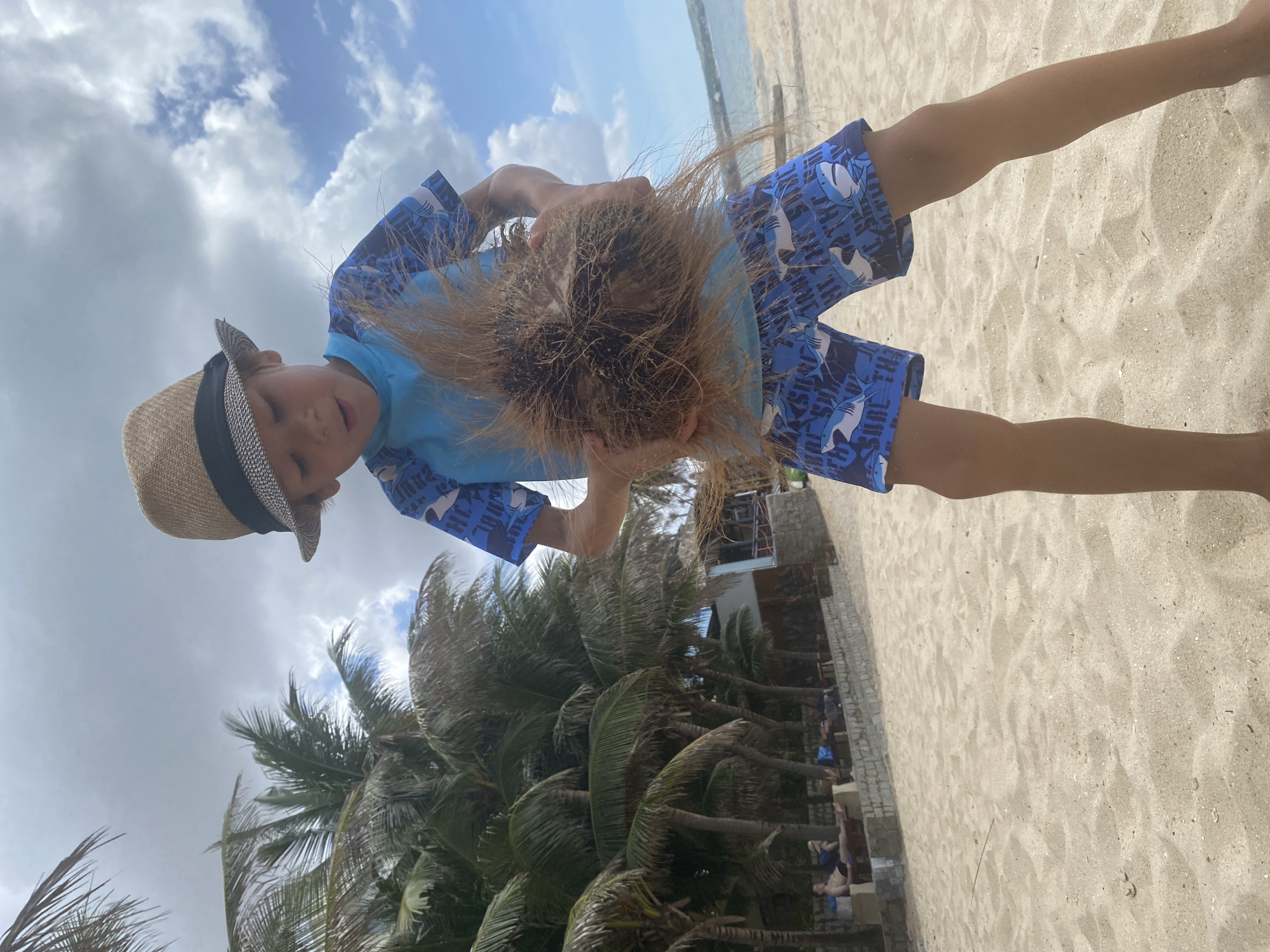 A young boy wearing a straw hat and blue shark-patterned swim trunks stands on a sandy beach. He is holding a large coconut husk, its fibrous exterior splayed outwards. The boy is looking down at the husk with an open mouth. In the background, palm trees line the edge of the beach, and beyond them, a few people are relaxing on loungers near a low stone wall. The sky is partly cloudy with patches of blue. The scene is consistent with a location in Mũi Né, Vietnam, known for its beaches and coconut groves.
