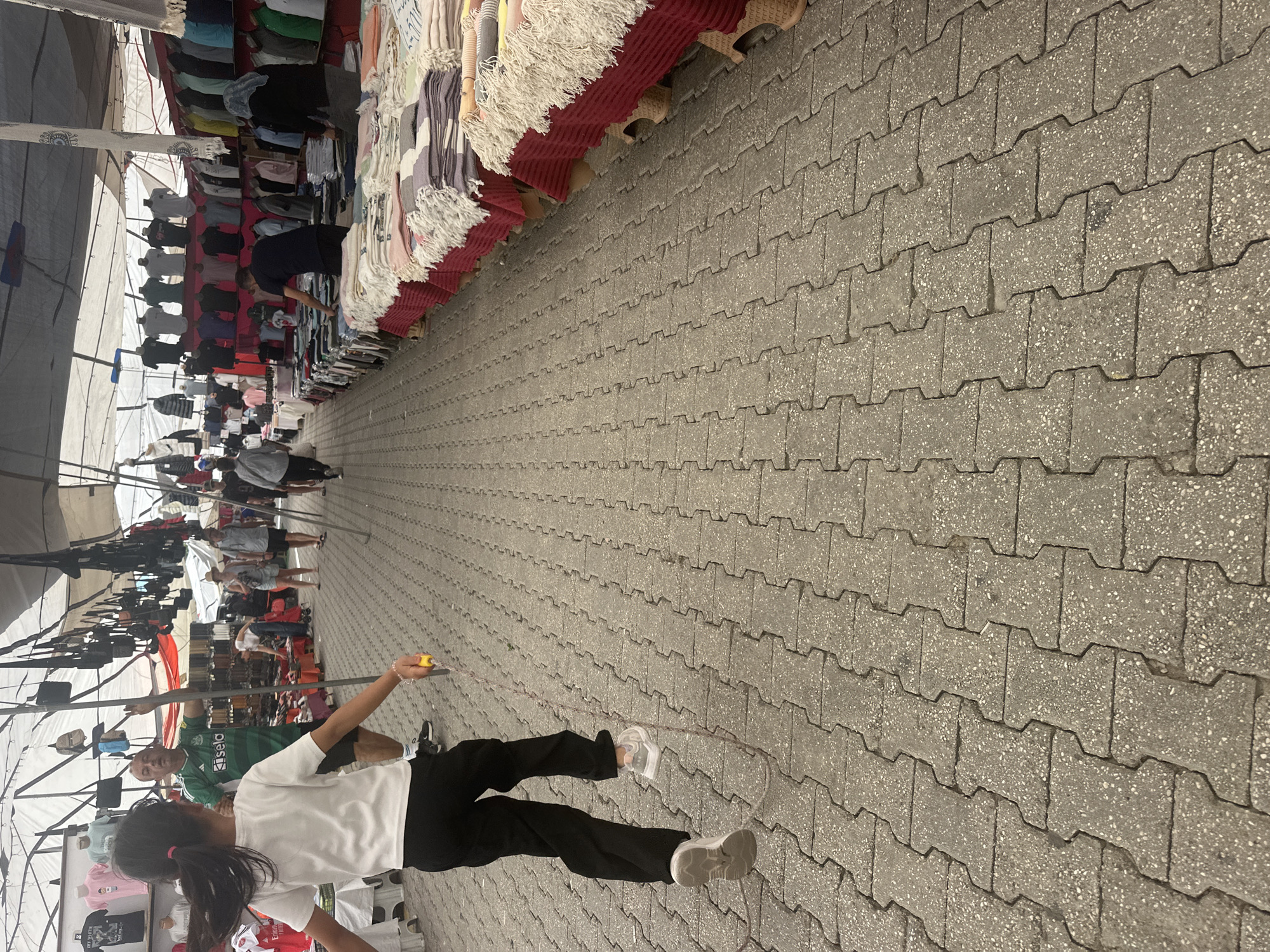 A girl with a ponytail and wearing a white shirt and black pants is jumping rope on a paved walkway in a bustling outdoor market. The location is likely Fethiye, Turkey, based on the prompt. The walkway is constructed of gray interlocking pavers, and vendors' stalls line both sides of the walkway. These stalls display various items, including clothing, scarves, and bags. Several people can be seen walking through the market in the distance. A man in a green soccer jersey stands to the girl's left. The scene is captured with an outdoor perspective, the image slightly tilted counter-clockwise.
