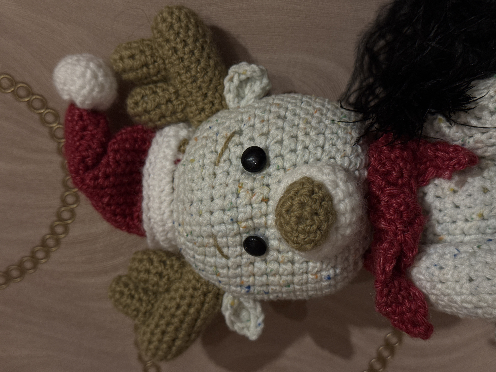 The media file displays a close-up, top-down view of a handcrafted, crocheted stuffed toy, likely an amigurumi, set against a wooden background. The toy appears to be a festive, perhaps Christmas-themed, character such as a reindeer or a bear dressed for the holidays.
The main body of the toy is made from light-colored (off-white or cream) yarn speckled with small, colorful flecks, giving it a tweed-like appearance. It has two small, round black safety eyes and a distinct, rounded brown crocheted nose. On the left side of its head, it wears a red crocheted Santa hat with a white pom-pom on the tip and a white band along the brim. Extending from the top of its head are two brown, multi-lobed crocheted elements, resembling antlers or ears. Around its neck or side, there is a vibrant red crocheted scarf. A section of fluffy, black material, possibly faux fur or feathers, is visible on the upper right side, suggesting hair or a mane.
The toy is positioned centrally in the frame. To the left, a metallic, possibly gold-colored, chain with circular links is visible, suggesting the toy might be part of a larger decoration or an ornament. The background is a warm-toned wooden surface, possibly a table or panel. No specific activity is taking place; the image is a static portrait of the crafted item. There is no visible text within the image. The scene is set indoors in Ново-Никольское, Russia.