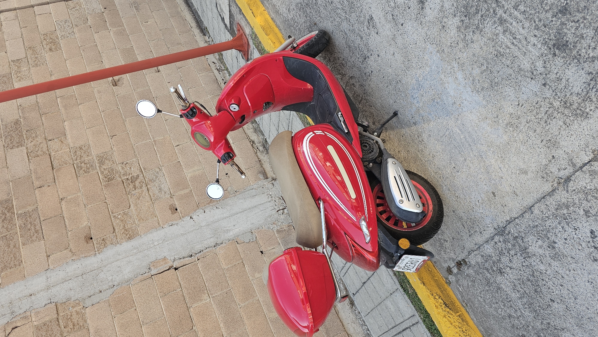 A red scooter is parked on a sidewalk in San Bernardino Tlaxcalancingo, Mexico. The scooter is angled slightly, with its front facing toward a building. The sidewalk is made of paving stones, and a red metal pole is visible nearby. A license plate is visible on the back of the scooter. The background consists of a concrete surface.