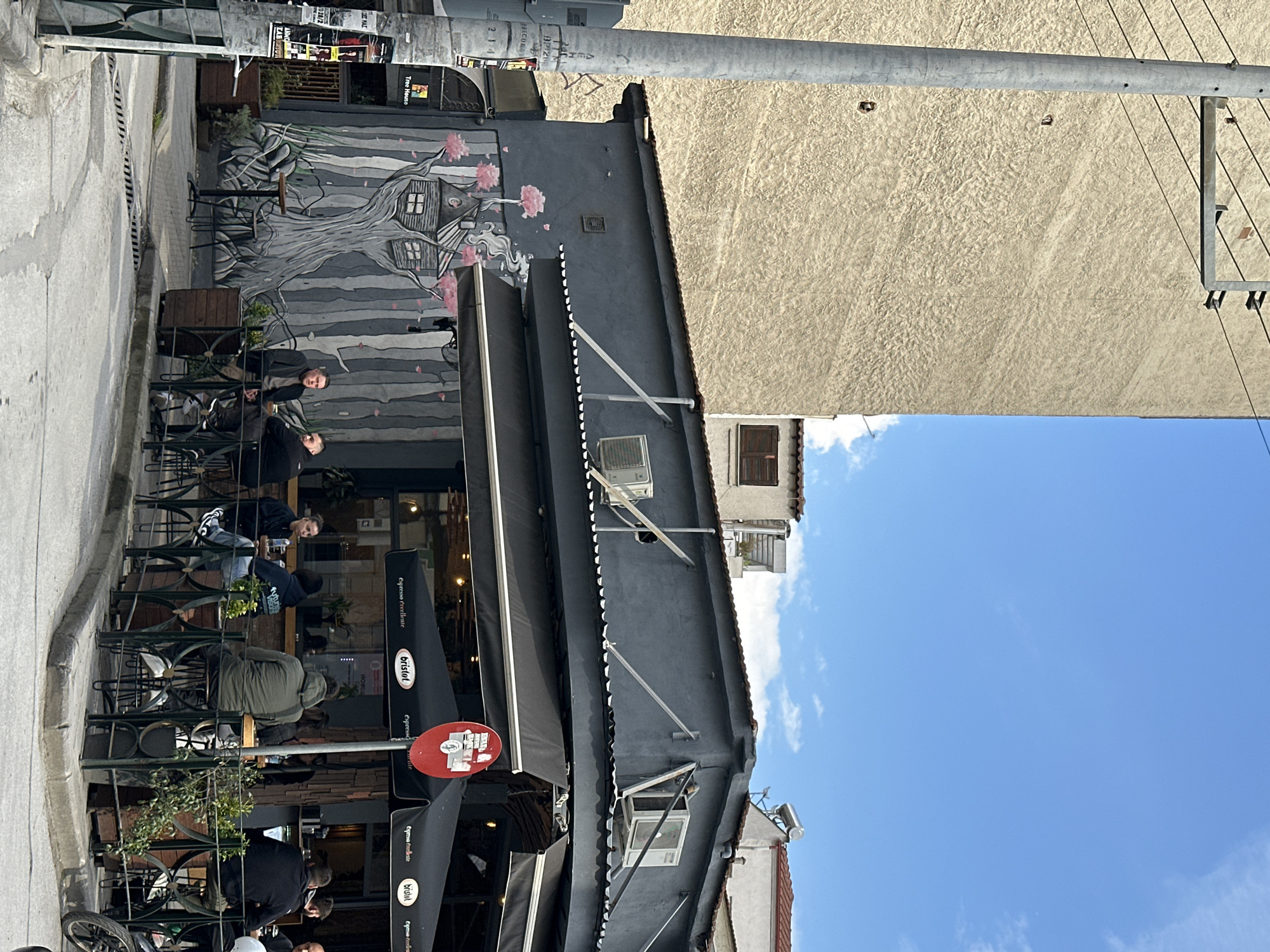 The image captures an outdoor dining scene in Serres, Greece. Several people are seated at tables under black awnings labeled "bristot espresso eccellente". A mural depicting a tree with house-like structures is visible on a building wall. A street sign with "Tree House" is visible. The sky is blue with scattered clouds. Buildings with light-colored facades are present in the background. A utility pole is in the upper part of the frame.