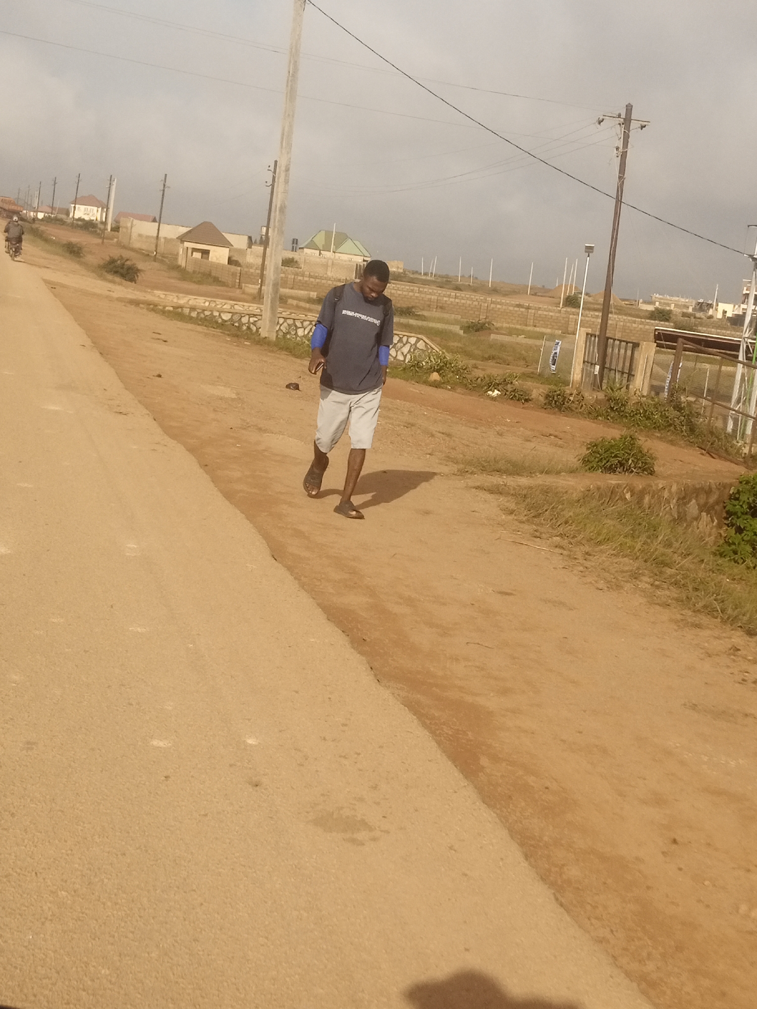 The image depicts a street scene in Kwang, Nigeria, under an overcast sky. A man is walking away from the camera on a dirt road, wearing a dark t-shirt with blue sleeves and light-colored shorts. He is also wearing dark sandals. To the left of the man, a paved road runs parallel to the dirt road. In the background, there are several buildings under construction, along with power poles and overhead wires. A motorcycle can be seen in the far left distance. The environment appears to be semi-urban with a mix of finished and unfinished structures.