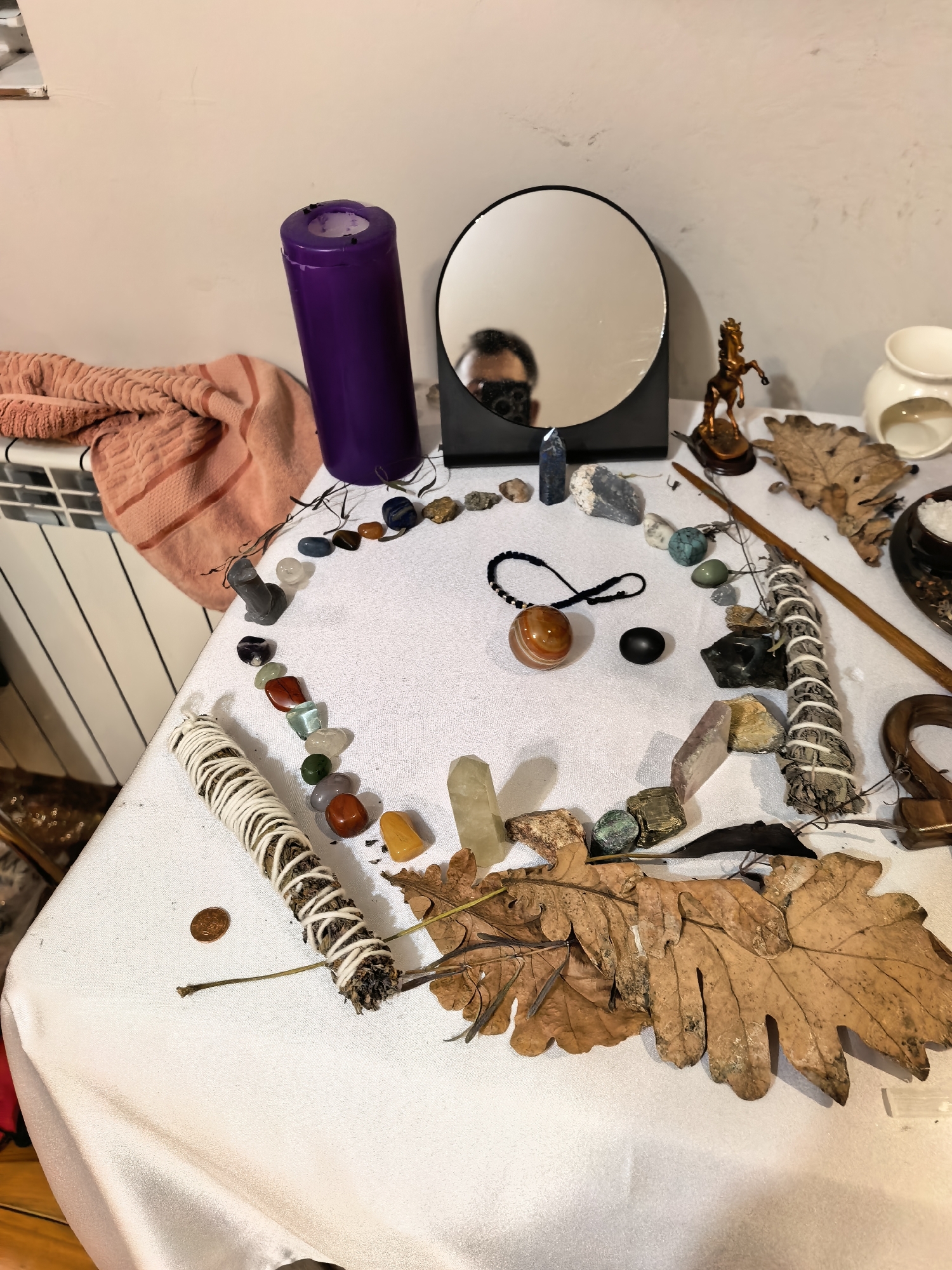 A white table with various objects arranged in a circular or semi-circular pattern sits indoors. The arrangement includes a purple candle, a round mirror reflecting a person holding a camera, various crystals and gemstones, a bronze horse figurine, a white ceramic oil warmer, dried leaves, a wooden wand, and bundles of sage. A peach-colored towel rests near a white radiator. The location is unknown.
