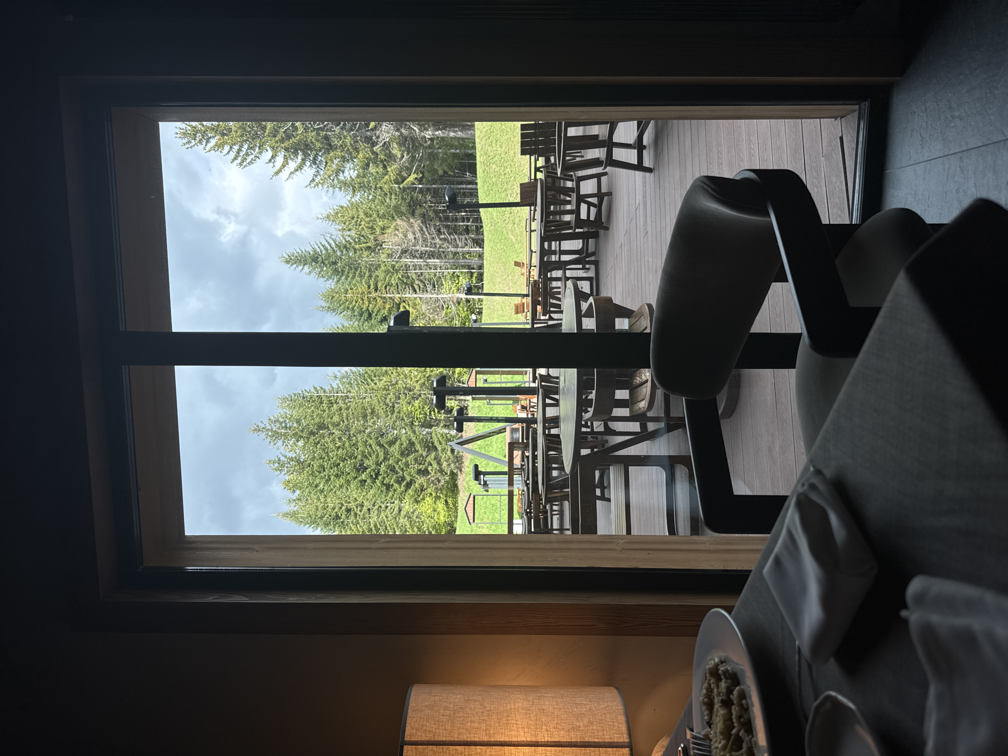 The image depicts a view from inside a building, possibly a restaurant, out onto an outdoor patio area. The view is framed by a large window or sliding glass door. Outside, there is a grassy lawn with several tall evergreen trees. A wooden deck is visible, furnished with wooden tables and chairs. Beyond the lawn, there are more trees in the background, and a cloudy sky is overhead. Inside, part of a dining table with a plate of food, a napkin, and a lamp are visible. The scene is in Katun Bačko brdo, Montenegro.
