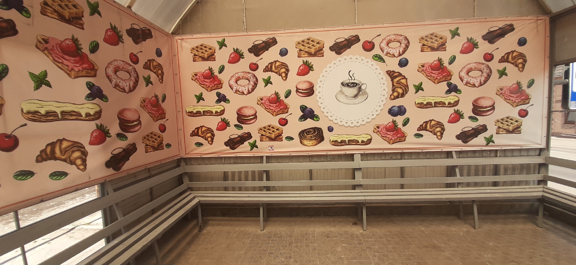The image shows a covered seating area, likely a bus stop or waiting shelter, with a large banner displaying various pastries and a cup of coffee. The banner is a pink background with a repeated pattern of illustrations of waffles, croissants, donuts, eclairs, macarons, strawberries, cherries, and blueberries. In the center of the banner is a circular decorative element with a drawing of a steaming cup of coffee on a saucer. The seating area has grey wooden benches that extend along two walls. The floor is paved with a checkered pattern. The overall impression is that of a quaint, sweet-themed waiting area. The banner and seating are likely located in Yessentukskaya, Russia, as indicated by the context. There are no people visible in the image, and the scene is static.