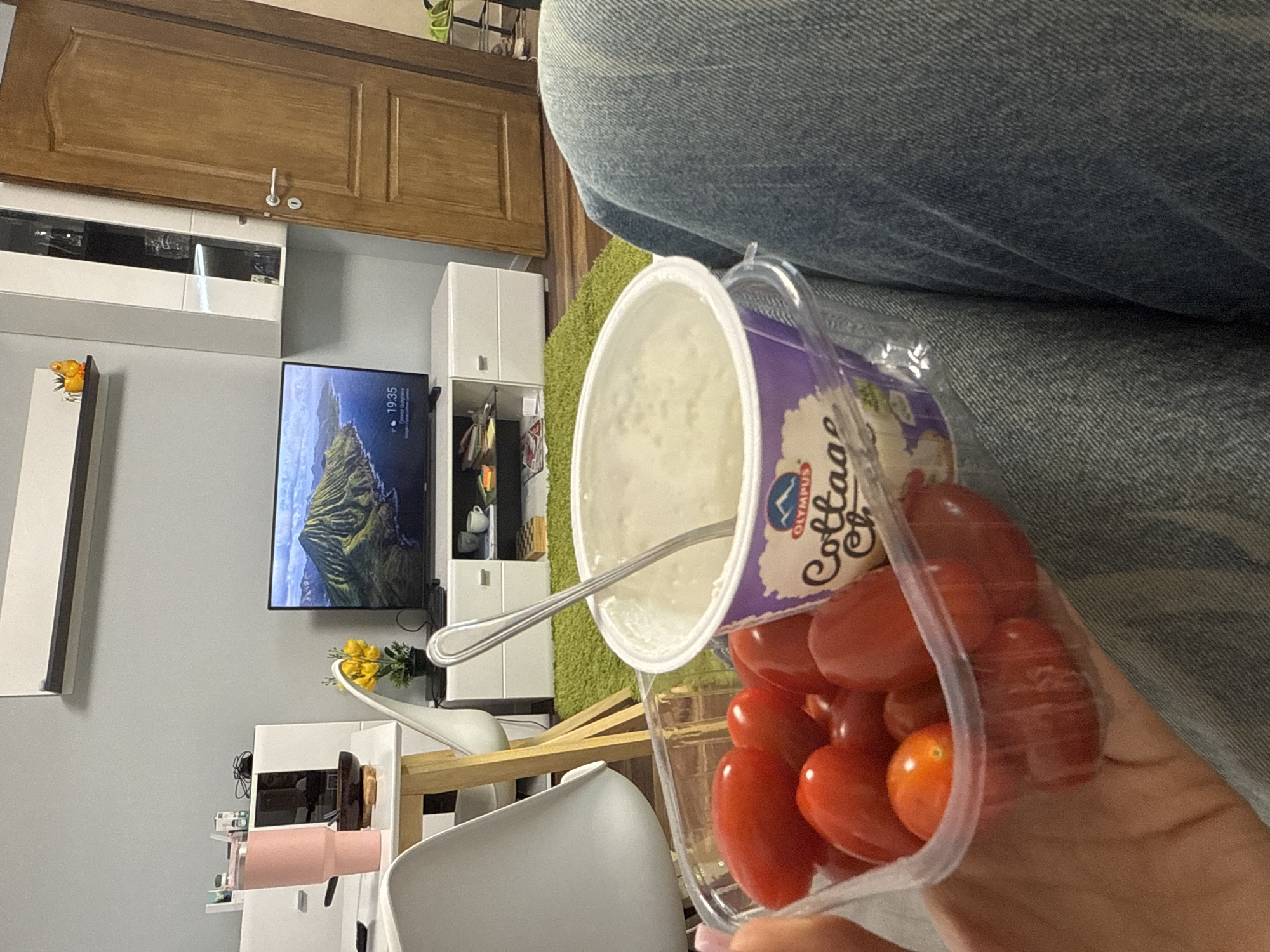 The image depicts a person sitting in what appears to be a living room. The person is holding a container of cherry tomatoes and a container of Olympus brand cottage cheese with a spoon in it. In the background, there is a television displaying a landscape scene, a white entertainment center, a wooden cabinet, and a white chair. The location is potentially in a home within the city of Sofia, Bulgaria, given the brand of cottage cheese available.