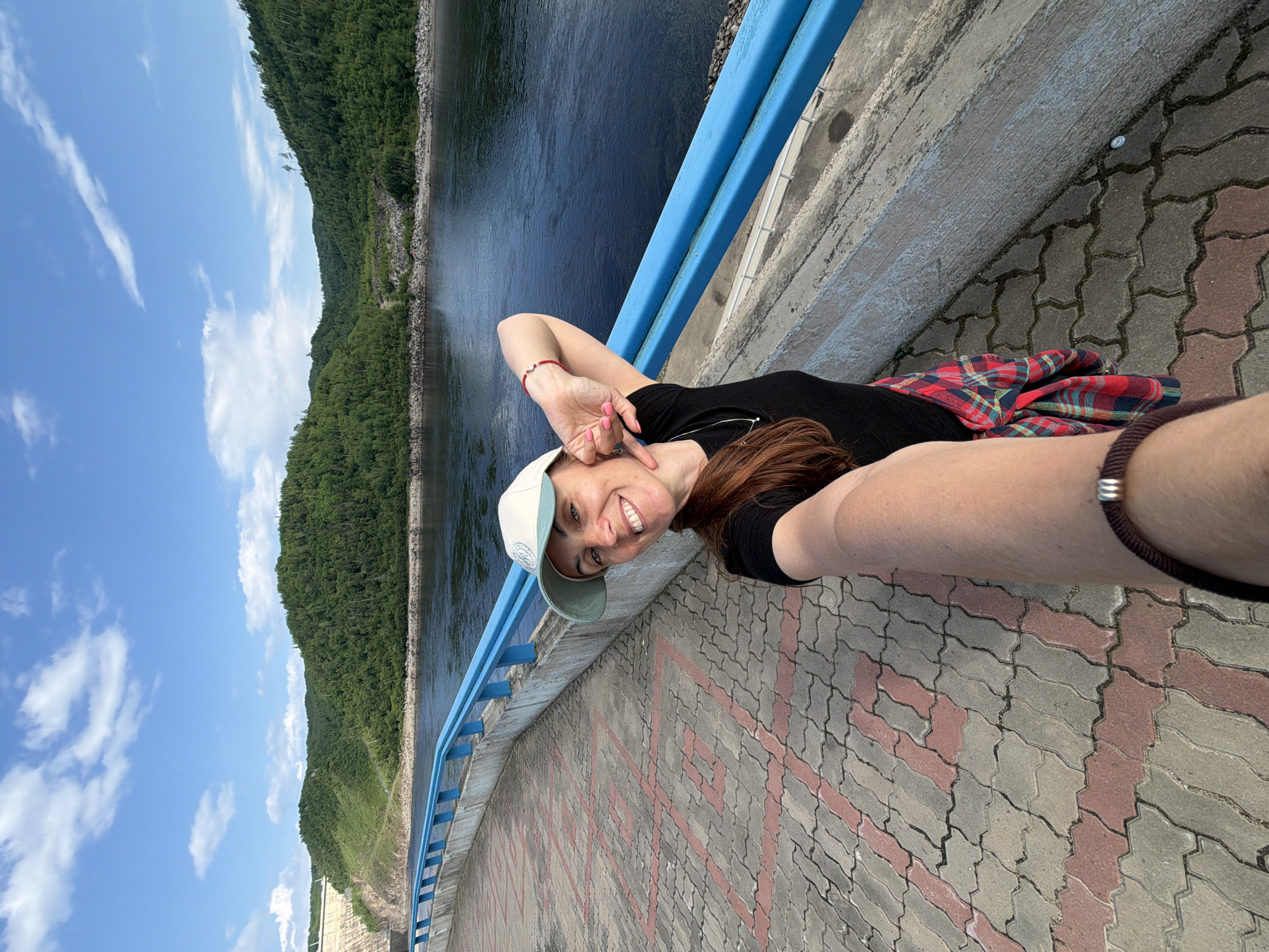 A woman is taking a selfie on a paved walkway next to a body of water and a forested landscape. The walkway is made of interlocking concrete pavers with a red pattern. A bright blue railing runs along the edge of the walkway. The woman is wearing a black t-shirt, a white baseball cap, and a red and black plaid shirt tied around her waist. She is smiling and has her left arm raised, with her hand near her face. The water in front of her is dark blue and calm, reflecting the sky. Beyond the water, a lush green forested hillside rises, with some bare patches and a road visible. In the far distance, a large concrete structure, possibly a dam, is partially visible on the left side of the frame. The sky is blue with scattered white clouds. The scene appears to be outdoors in a natural setting, likely a recreational area near a body of water. The location is identified as the Talakan Dam in Talakan, Russia.