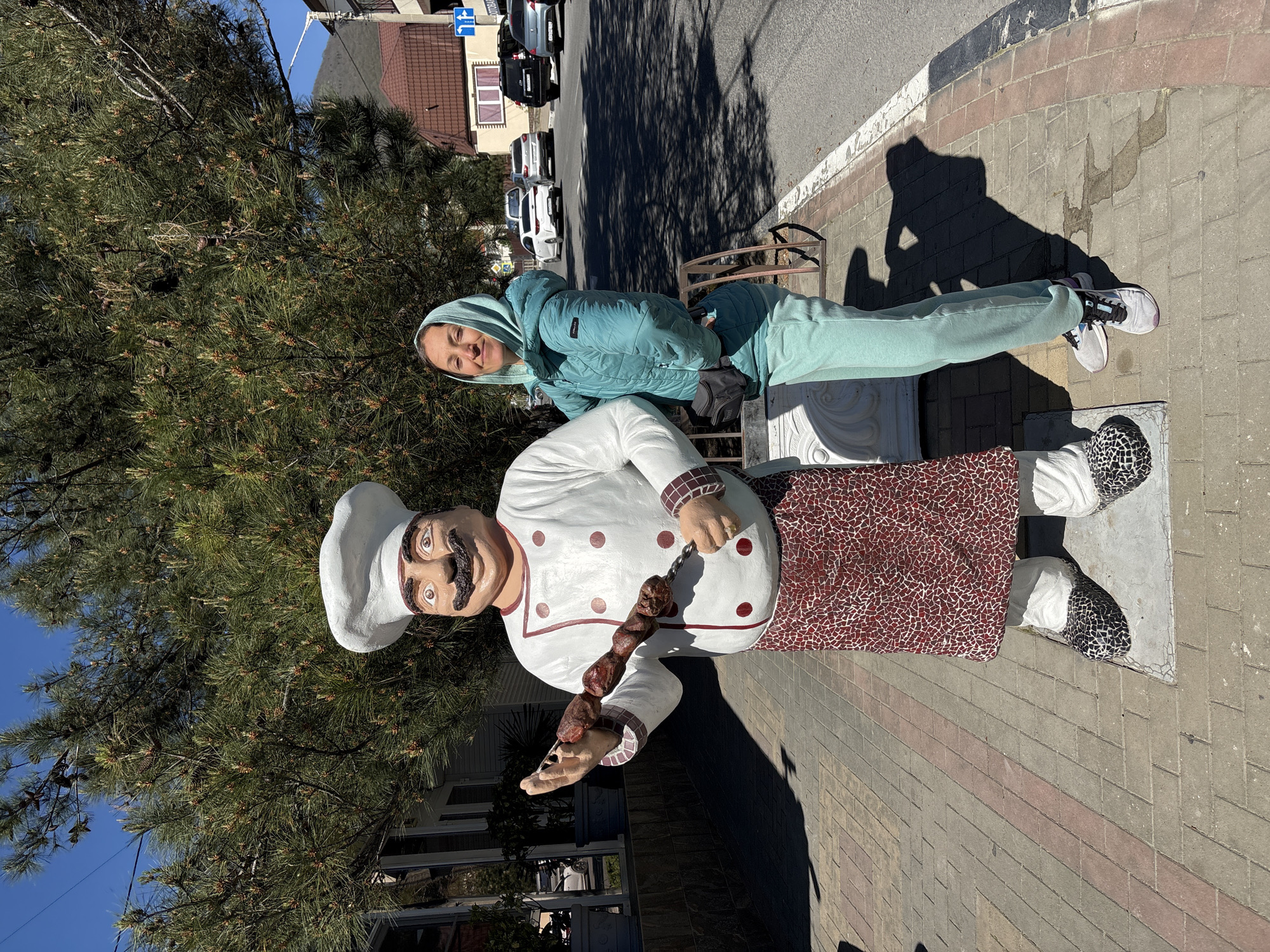 This image captures a woman posing outdoors next to a large chef statue in Gelendzhik, Russia, under bright sunshine. The woman, smiling towards the camera, wears a light teal hooded puffer jacket, matching sweatpants, and white sneakers, with a dark fanny pack across her chest.
The prominent chef statue is depicted in a white chef's hat and coat with red polka dots and checkered trim, a checkered reddish-brown and white apron, and has a large brown mustache. It holds a metal skewer laden with pieces of meat, resembling shish kebab or shashlik. The statue's base features patterned, possibly mosaic, elements.
The scene is set on a paved sidewalk next to a large pine tree. In the background, a street with several parked cars and residential buildings under a clear blue sky is visible. Strong sunlight casts distinct shadows of the subjects and trees onto the pavement, indicating a clear, bright day. A blue road sign with white arrows (one pointing left, another downward-left) is visible above the parked cars.