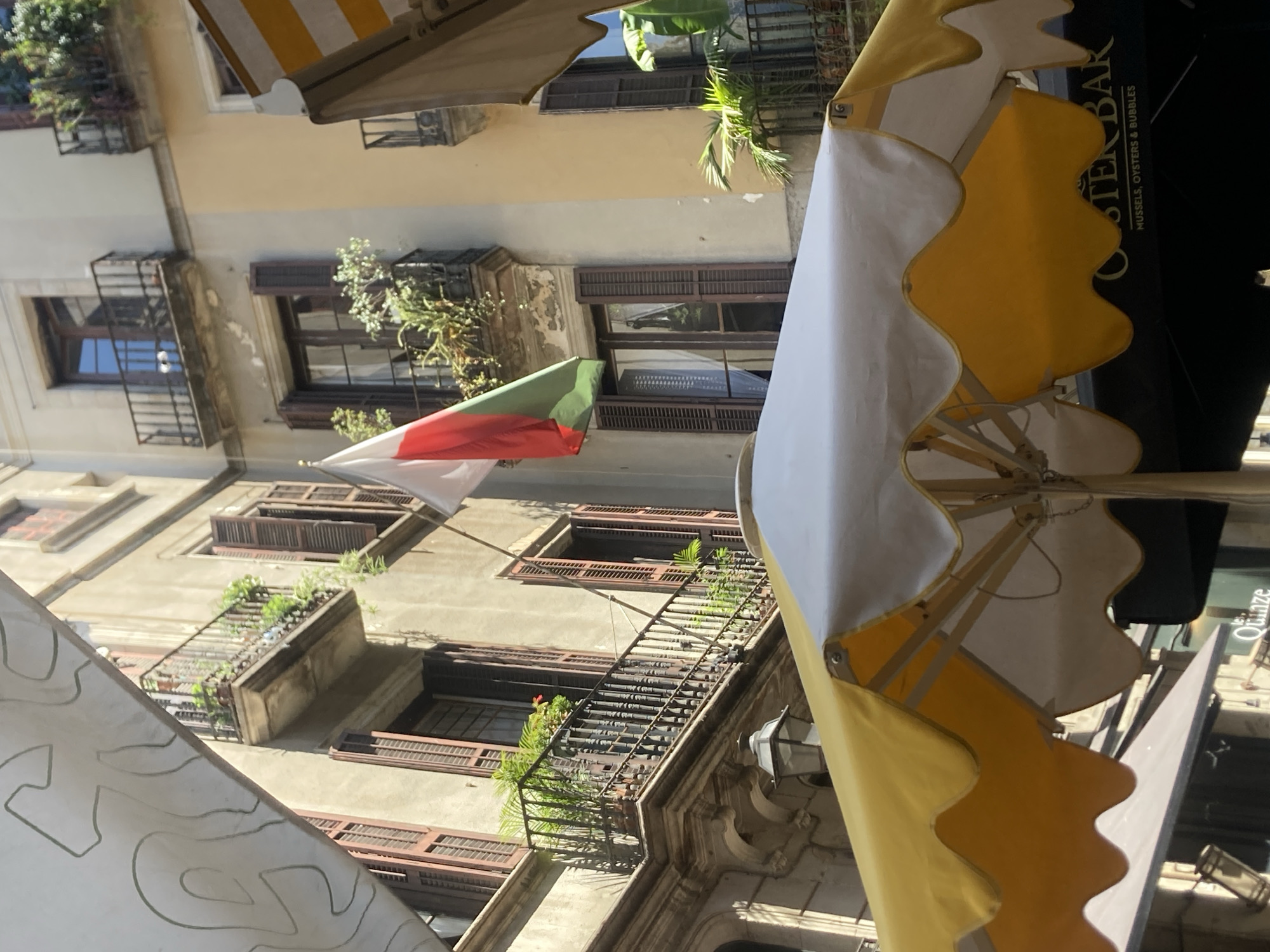 The image depicts a narrow street in Barcelona, Spain, with a focus on the architecture of residential buildings and a foreground element of outdoor dining umbrellas. A flag, appearing to be the flag of Bulgaria, hangs from one of the buildings, featuring three horizontal stripes of white, green, and red. The buildings themselves are characterized by traditional Catalan architecture, with numerous balconies adorned with potted plants and ornate railings. Many windows feature wooden shutters, some of which are open, revealing glimpses of the interiors. In the foreground, a large umbrella with yellow and white scalloped edges dominates the lower portion of the frame, casting a shadow and suggesting a dining area for a cafe or restaurant. Below the umbrella, a sign for "Oyster Bar" with the tagline "Mussels, Oysters & Bubbles" is partially visible, indicating the establishment's offerings. The scene conveys a sense of a vibrant, sun-drenched street in a European city, likely a popular tourist destination given the presence of outdoor dining and the flag suggesting international presence. No people are clearly visible in the frame, but the setup implies a daytime scene with patrons potentially seated at the outdoor tables.