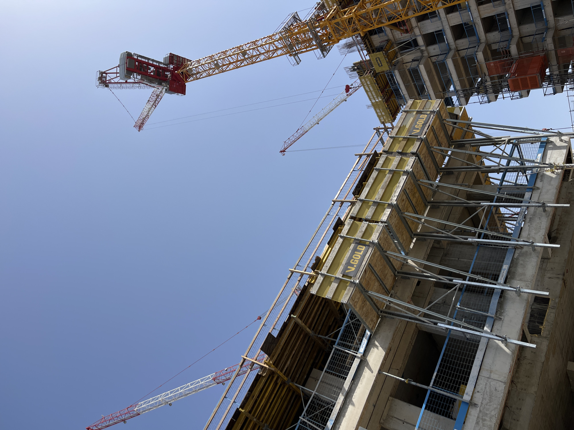 The image captures a low-angle view of a construction site in Tel-Aviv, Israel, under a clear, light blue sky. The primary subjects are the building under construction, several cranes, and the scaffolding and support structures used in the building process.
The building's structure is visible, revealing its skeletal frame with concrete and metal reinforcements. Scaffolding, marked with "V.GOLD" in yellow lettering, is attached to the building, suggesting ongoing work.
Three cranes are visible in the shot, each with a distinct color scheme. One is primarily yellow, with a red and white jib crane further in the background. The third is a white and red jib crane located closer to the bottom left corner of the image.
The environment is urban, as suggested by the construction and the likelihood of this type of construction in a city like Tel-Aviv. The absence of people indicates that the photo was taken without immediate human presence in the visible frame, focusing instead on the architectural and engineering aspects of the site.